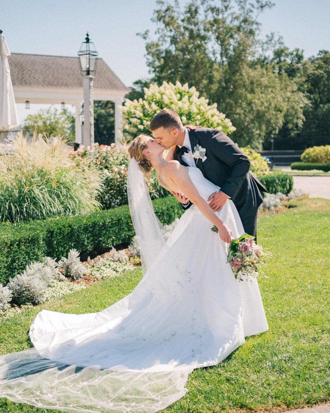 A bride in a white wedding dress and veil is being lifted by a man in a dark suit, both standing on a grassy area with a garden and a white gazebo…