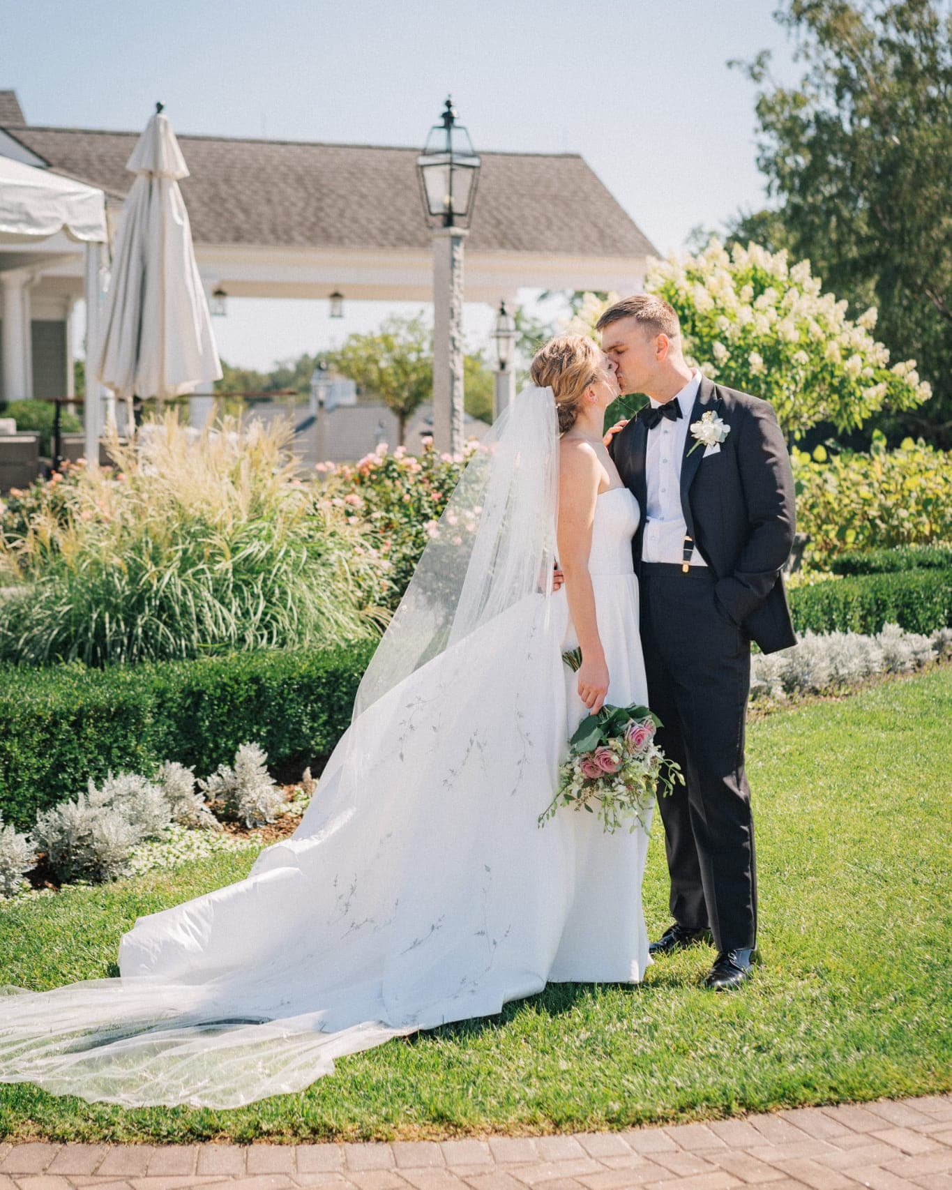 A bride in a white gown and veil kisses a groom in a black suit, standing on a grassy lawn with a garden and building in the background.