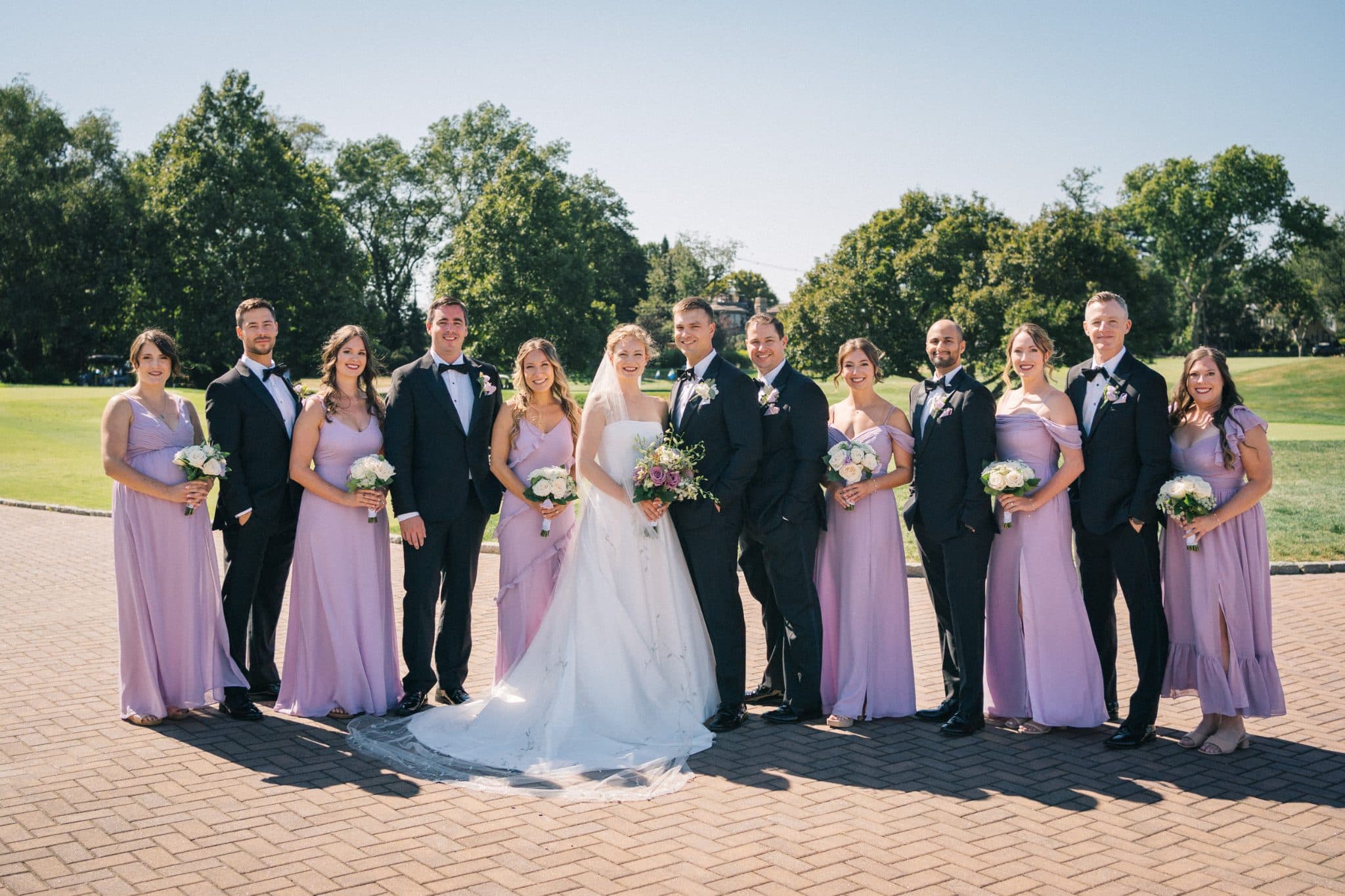 A wedding party stands together outdoors, with bridesmaids in lavender dresses and groomsmen in black suits, holding bouquets.