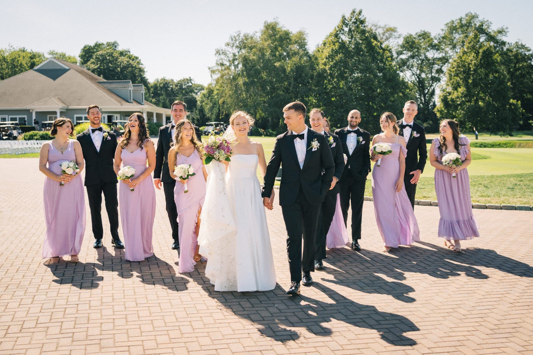 A wedding party in formal attire walks together on a brick path, holding bouquets.