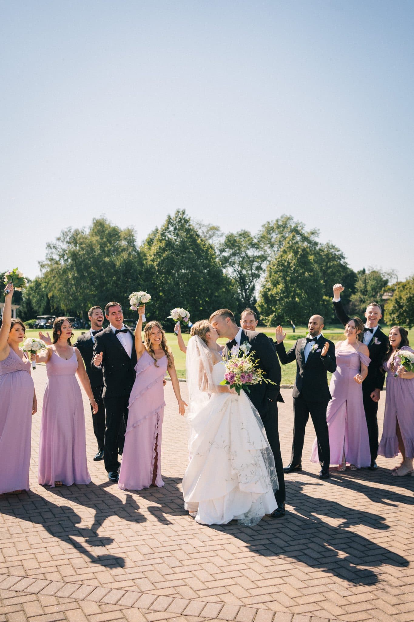 A bride in a white gown and groom in a black suit kiss, surrounded by bridesmaids in lavender dresses and groomsmen in black suits, under a clear sky.