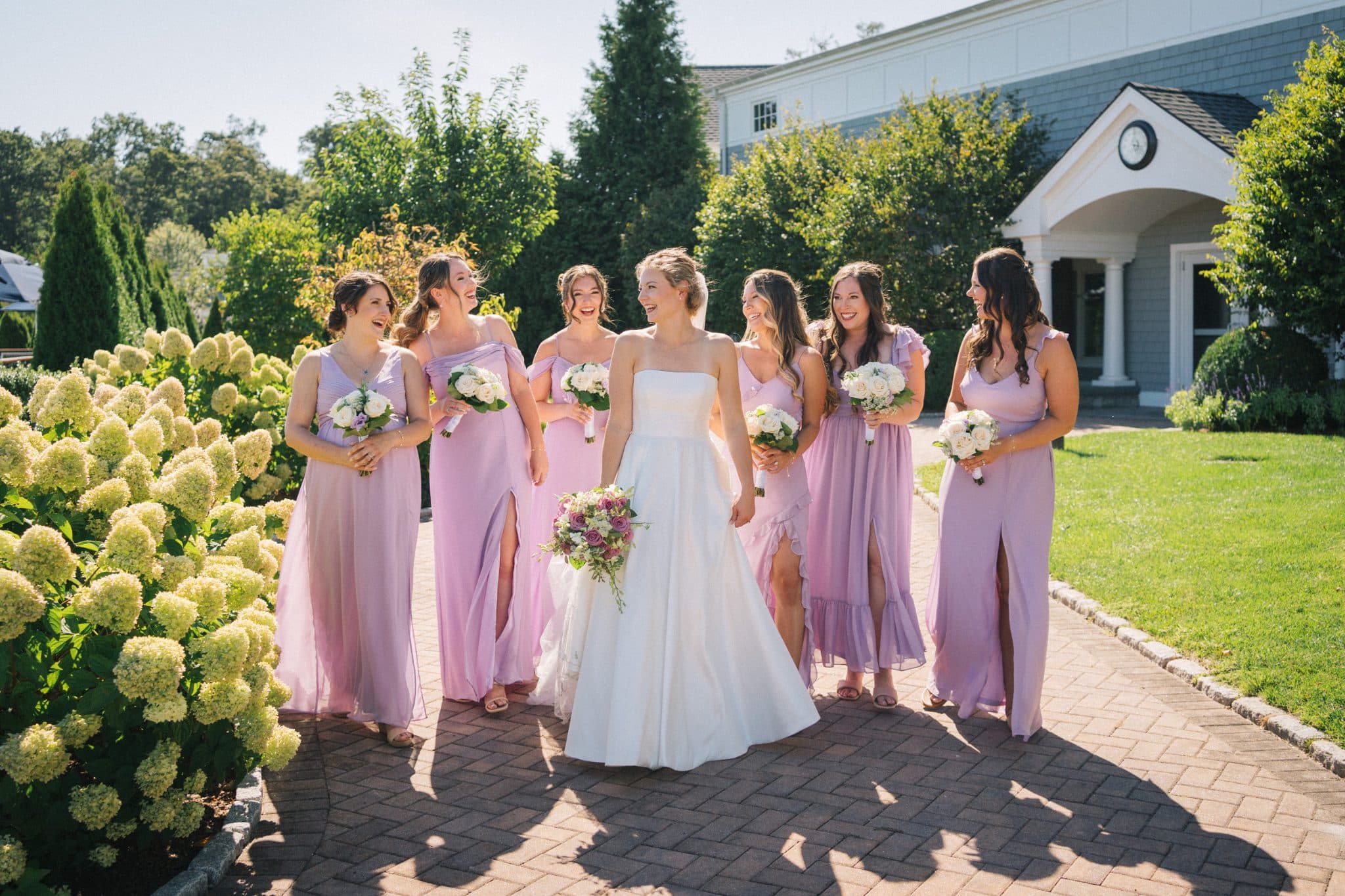 A bride in a white dress walks with bridesmaids in lavender gowns, holding bouquets, on a brick path near a building and greenery.