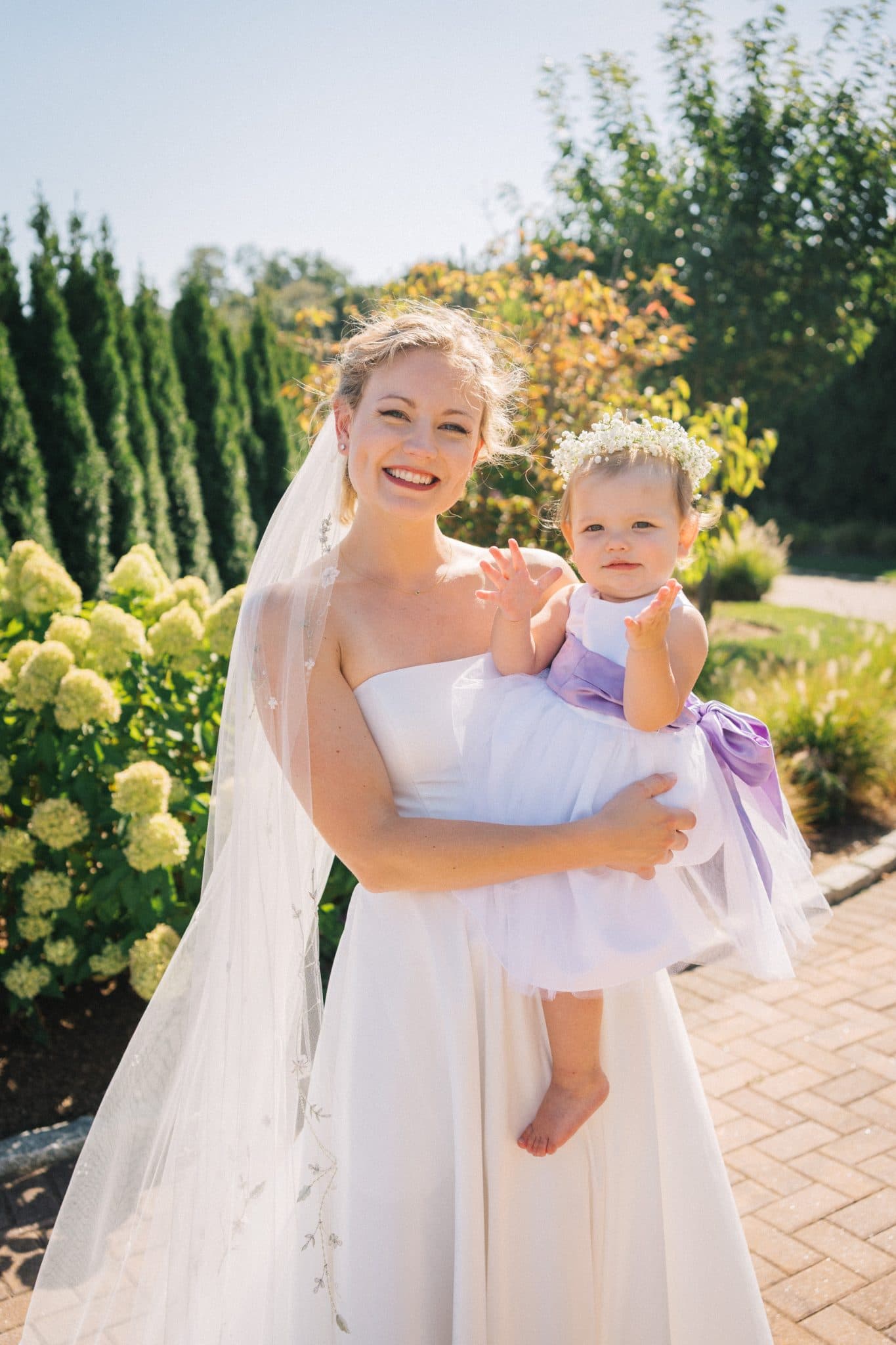 A bride in a white wedding dress holds a baby wrapped in a white and purple cloth, surrounded by greenery and a brick path.