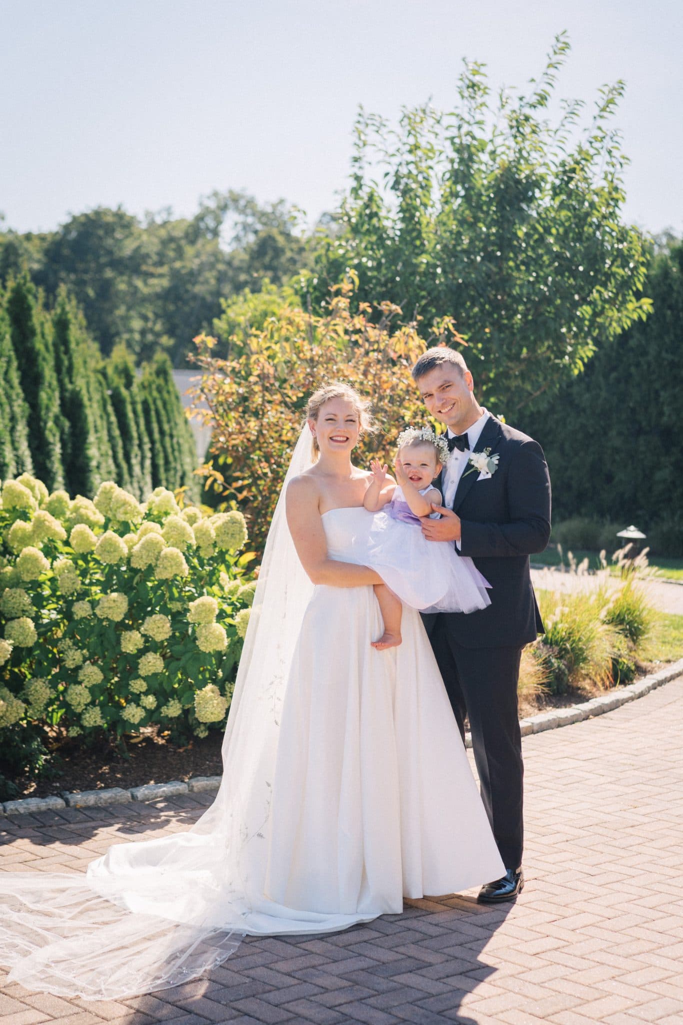 A bride in a white gown and veil holds a child in a purple dress, standing next to a groom in a dark suit with a bow tie, in a garden setting.