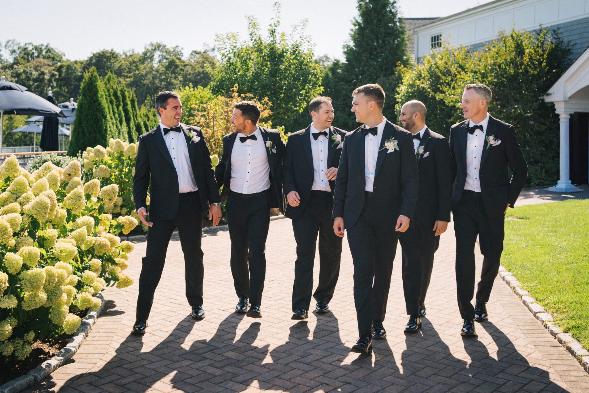 Six men in black tuxedos walk together on a brick path near a building and greenery.