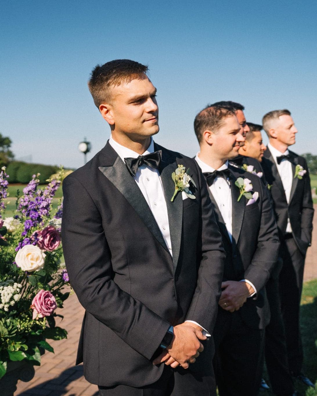A groom and his groomsmen in tuxedos stand in a garden with a floral arrangement in the foreground.
