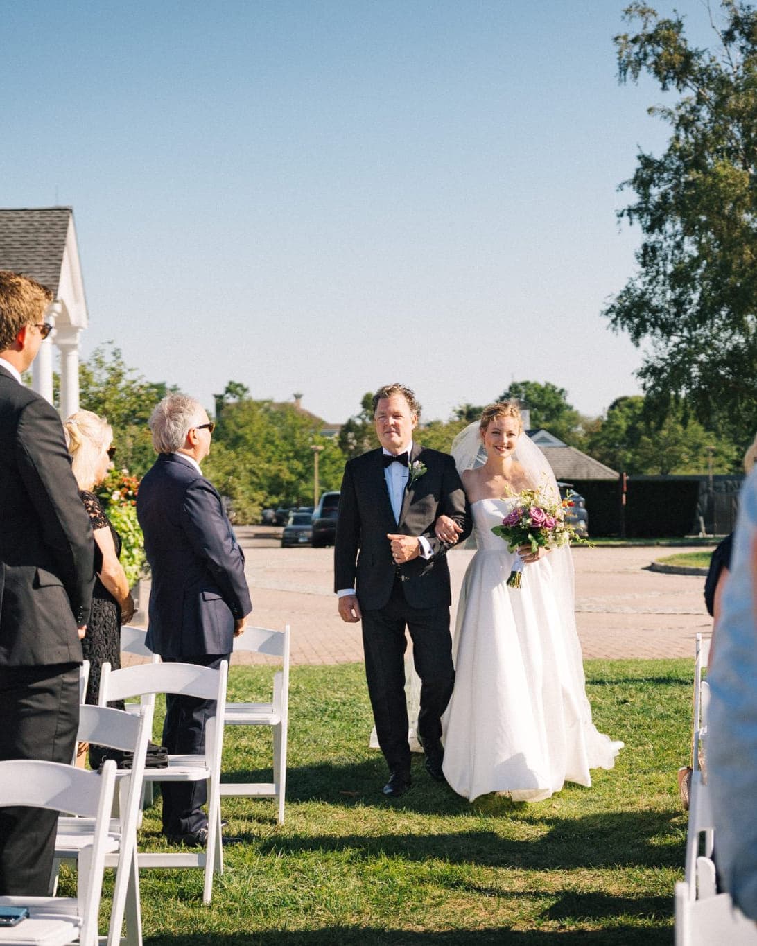 A bride and her father walk down a grass aisle during an outdoor wedding ceremony.