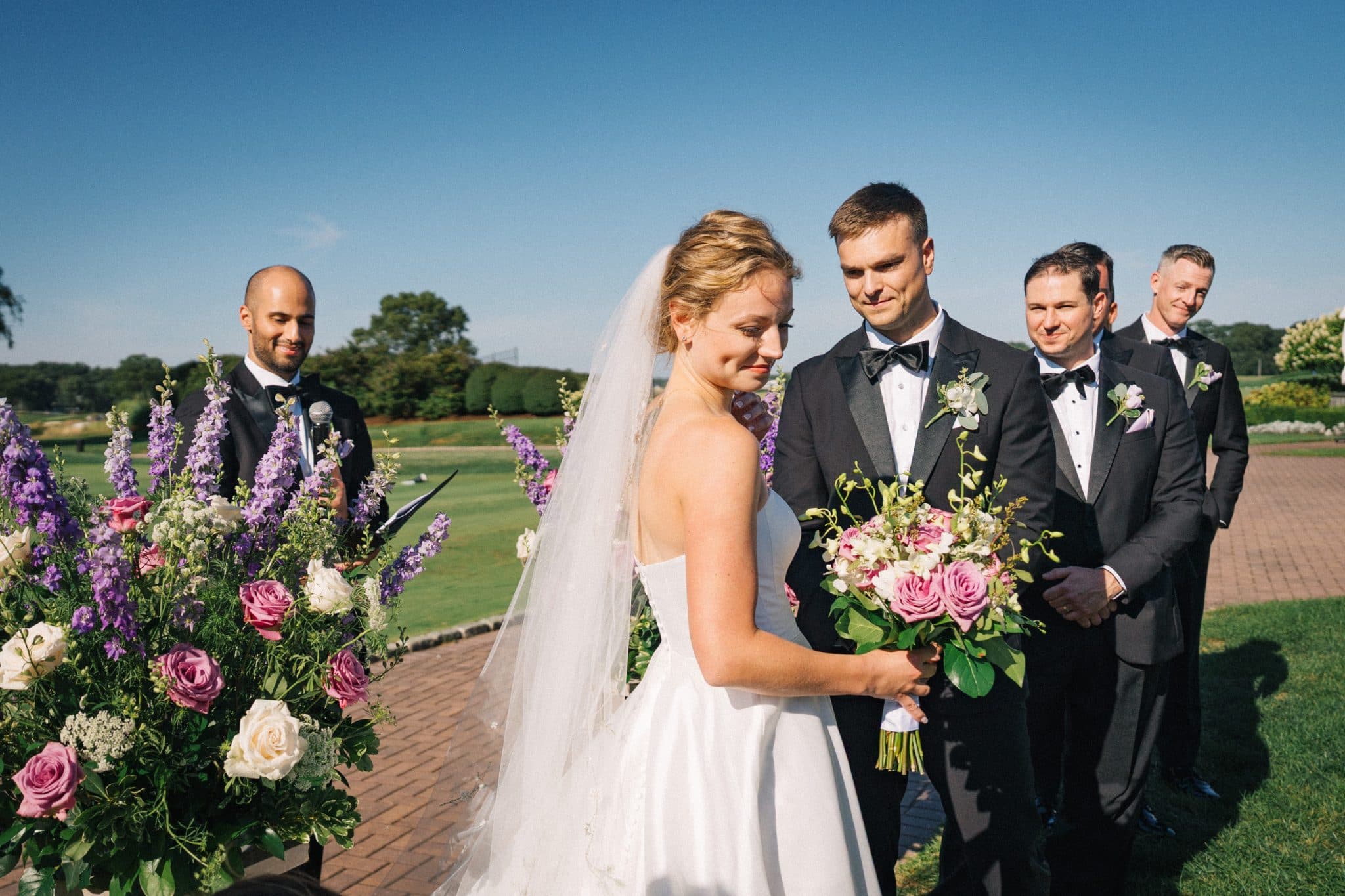 A bride in a white dress holds a bouquet, standing with groomsmen in black suits and bow ties under a clear blue sky.
