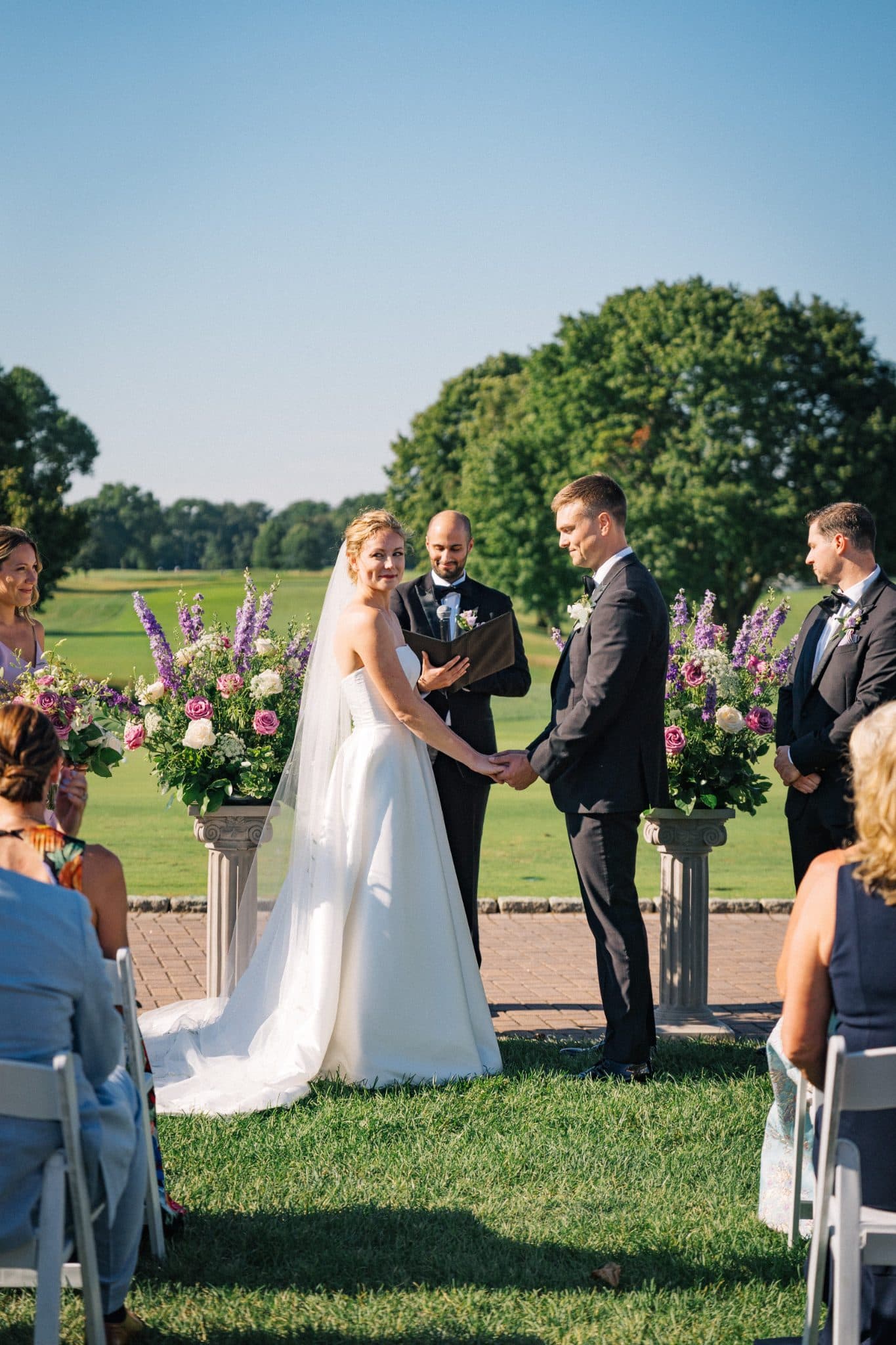 A bride in a white gown and a groom in a dark suit hold hands during an outdoor wedding ceremony with guests seated and floral arrangements.