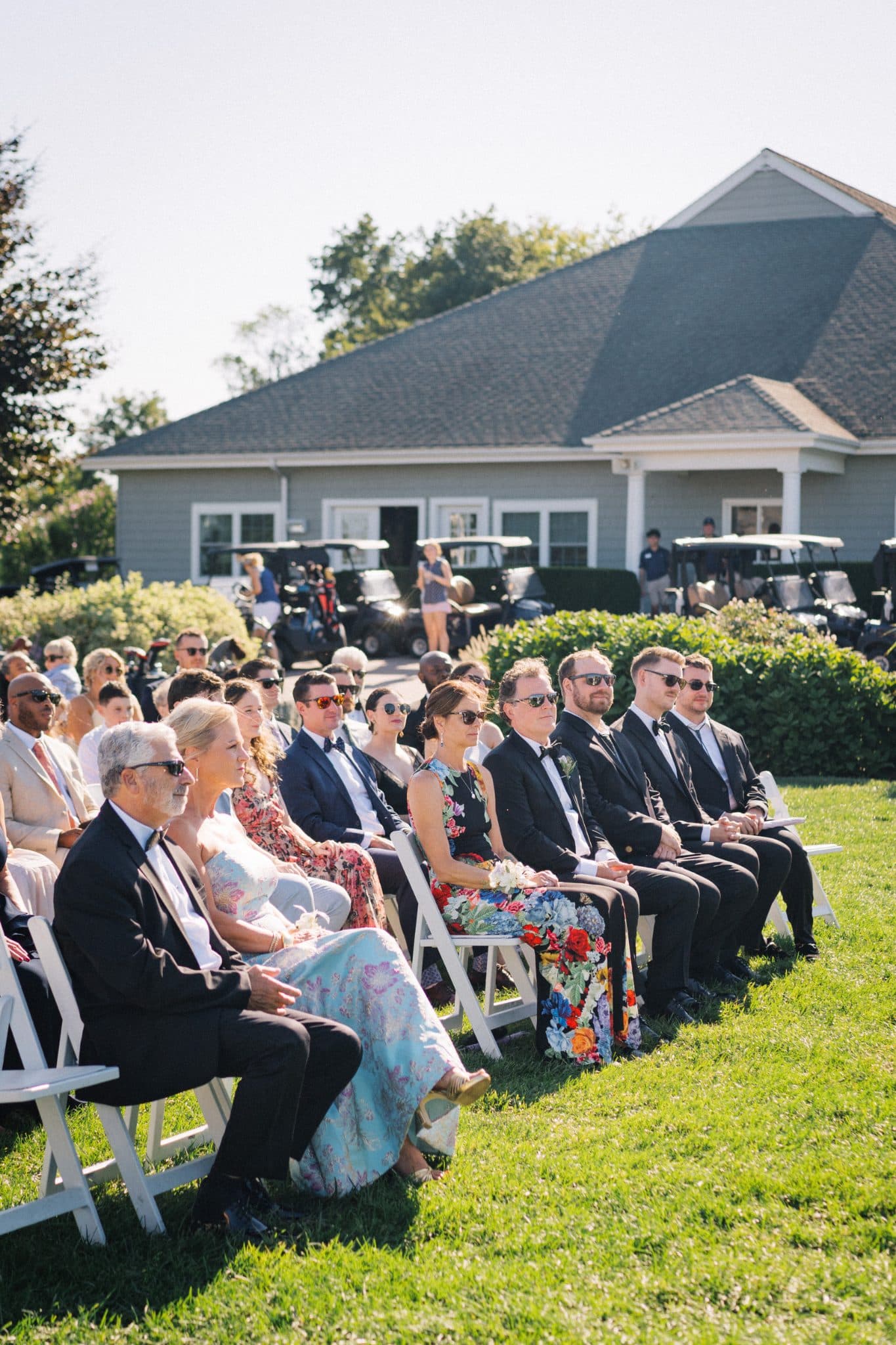 A wedding ceremony scene with guests seated on white chairs in front of a building with golf carts in the background.