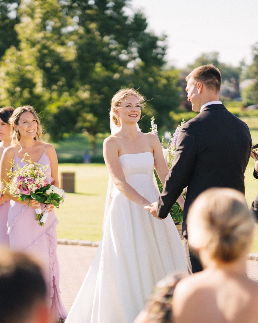 A bride in a white dress stands at the altar, holding hands with a groom in a black suit, surrounded by bridesmaids in lavender dresses and guests.
