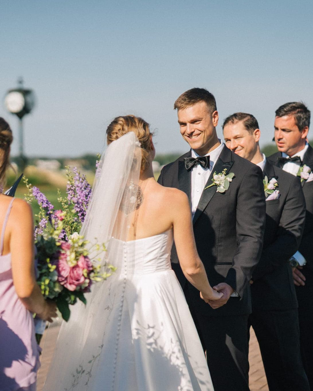 A bride and groom stand facing each other, surrounded by bridesmaids and groomsmen in formal attire, holding bouquets of flowers.