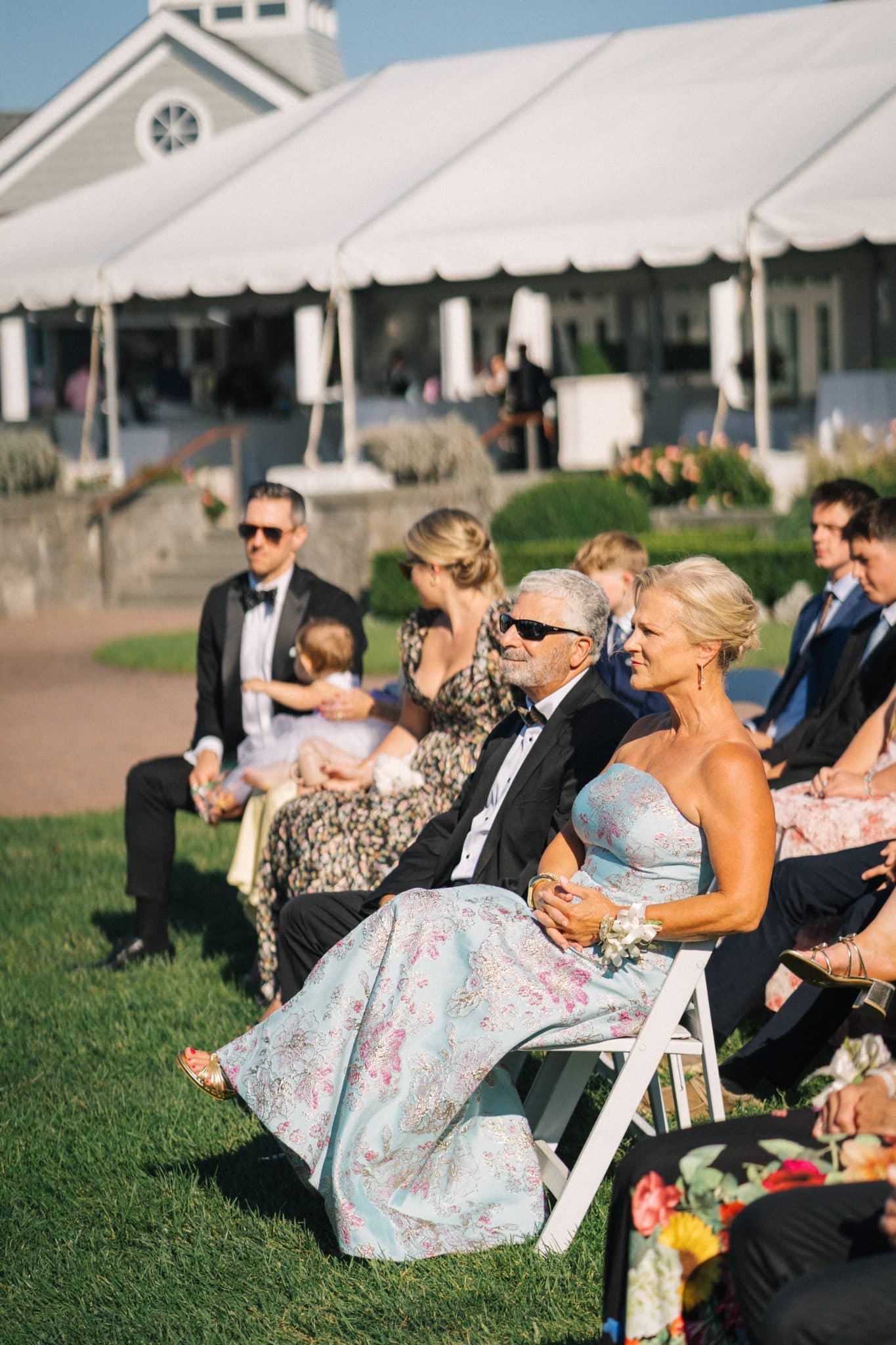 A group of people in formal attire sits on chairs under a white tent, watching a ceremony on a grassy lawn.