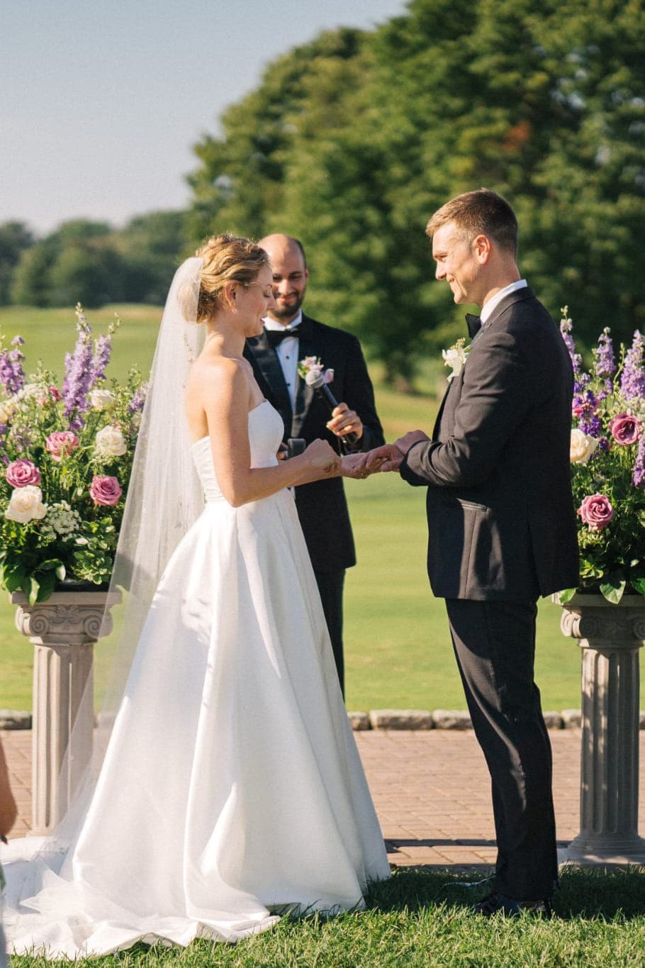 A bride and groom exchange rings during an outdoor wedding ceremony with guests and floral arrangements in the background.