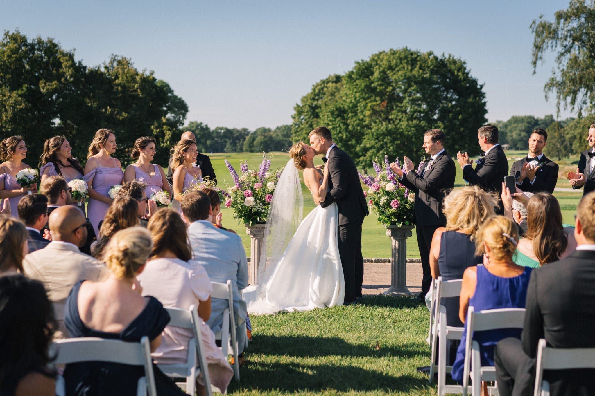 A bride and groom share a kiss at their wedding ceremony, surrounded by guests and floral arrangements.
