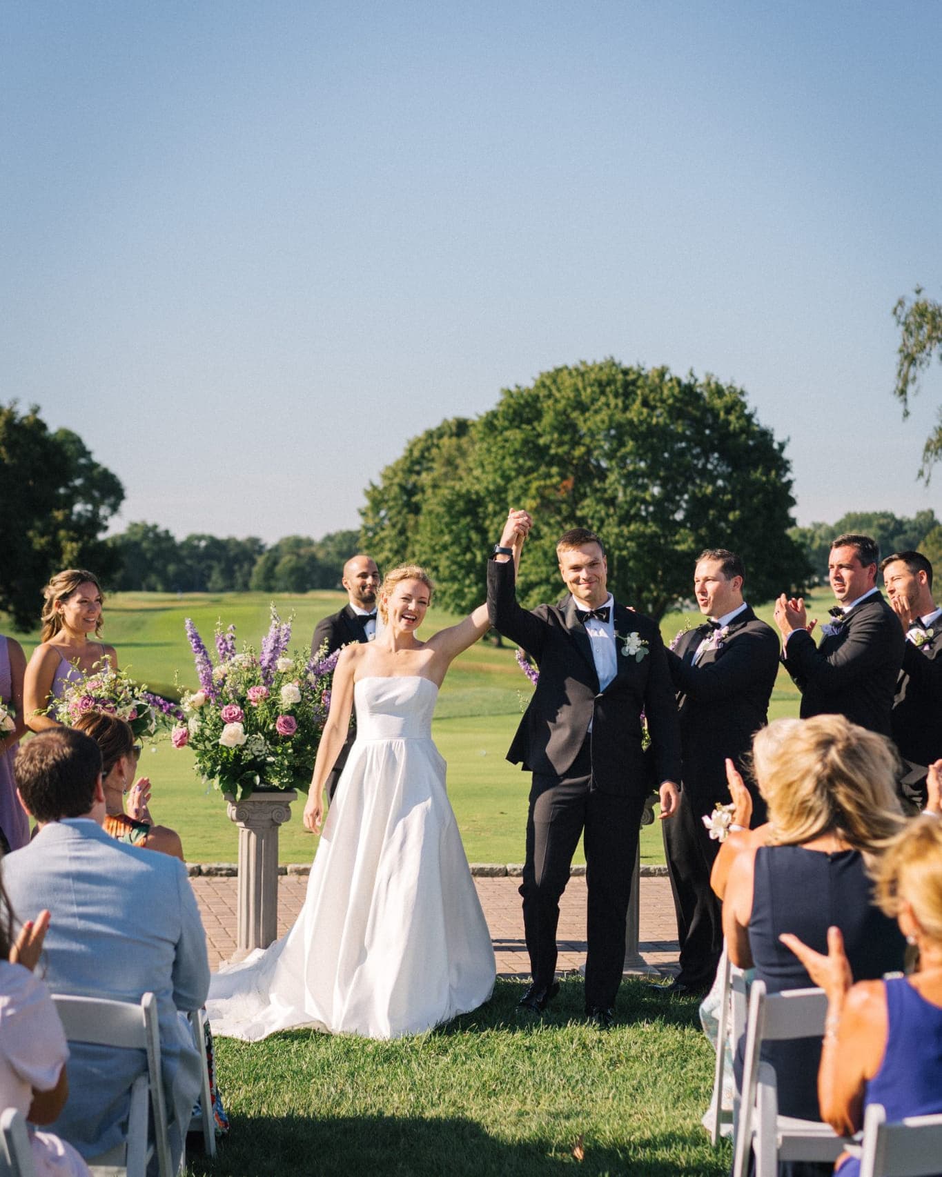 A bride and groom hold hands, celebrating at an outdoor wedding ceremony with guests clapping and flowers in the background.