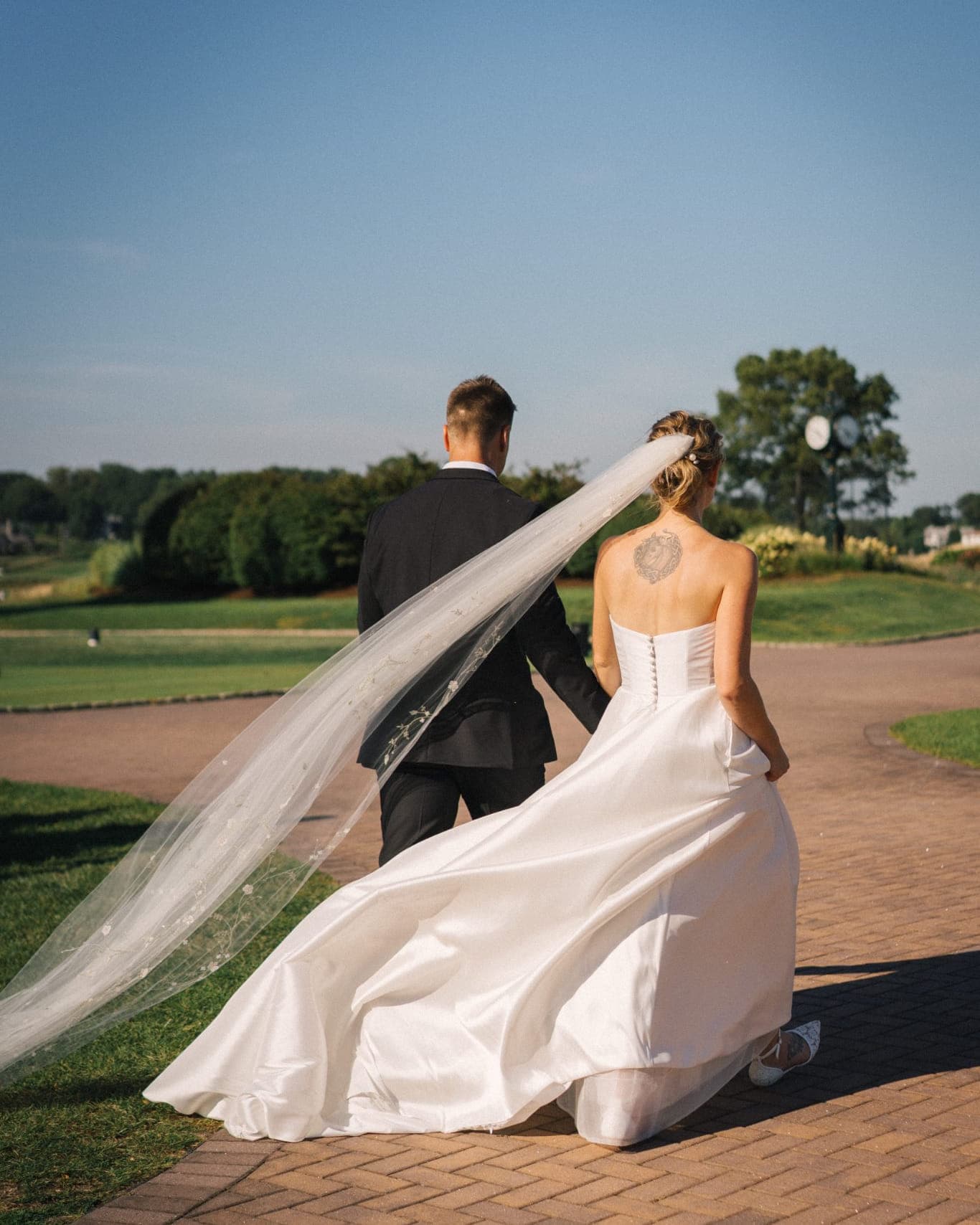 A bride and groom walk hand in hand on a brick path, the bride's veil flowing behind her.