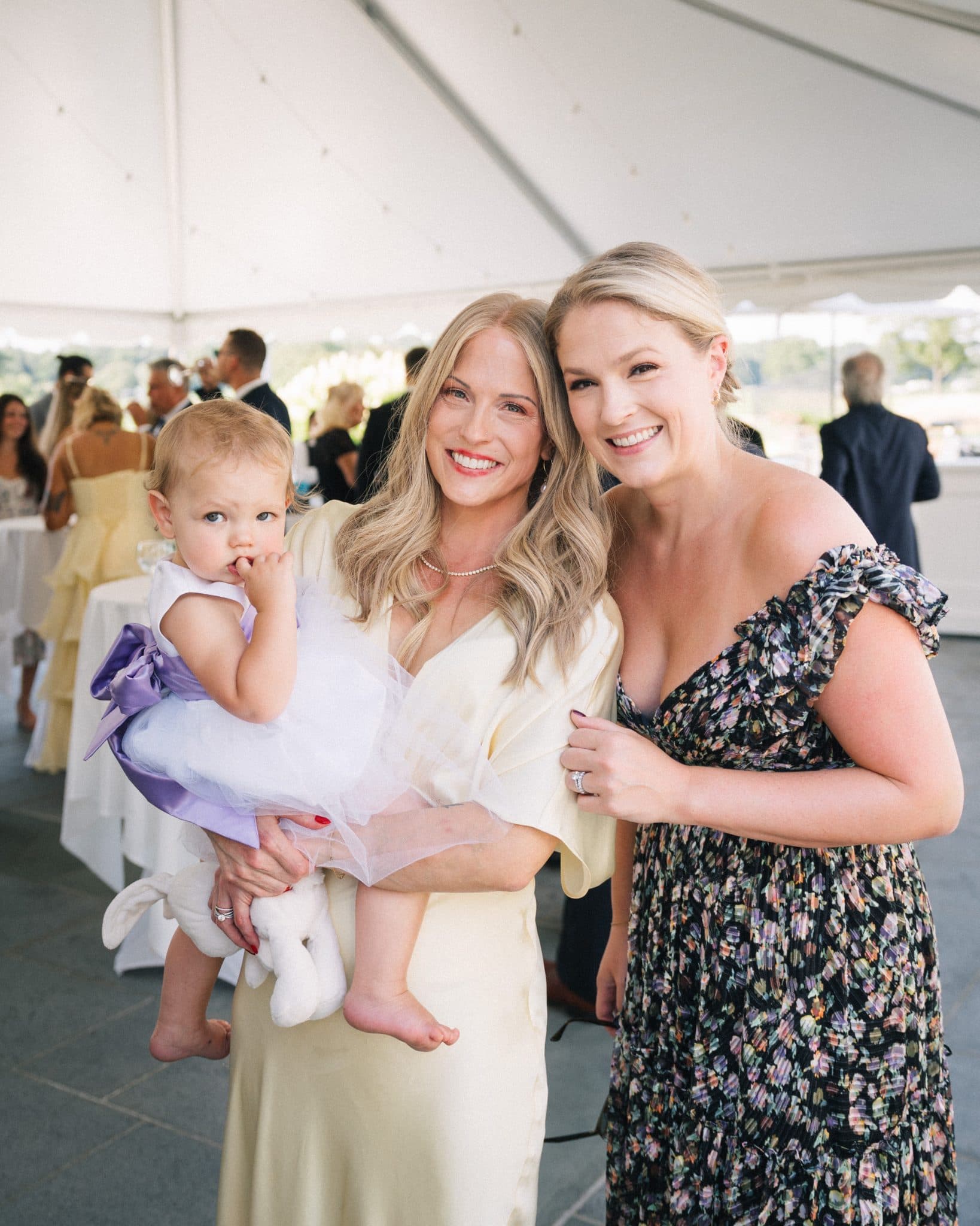 Two women smile, one holding a baby dressed in white with a purple bow, under a tent at a social gathering.