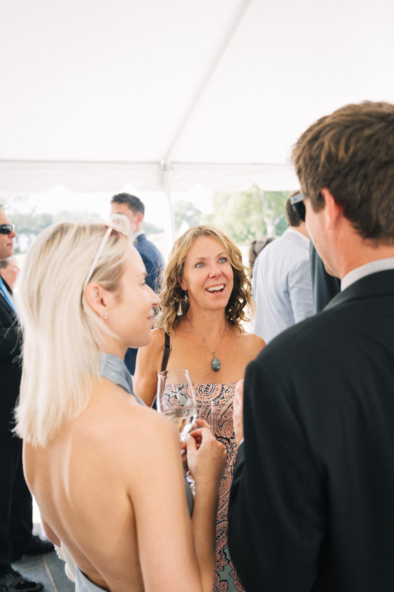 A woman in a patterned dress smiles while holding a wine glass, surrounded by other people at an outdoor event under a tent.