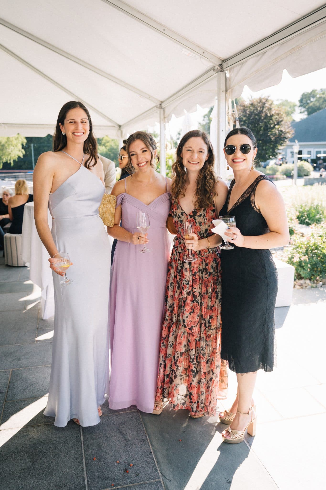 Four women in dresses stand under a tent, holding drinks, smiling at the camera.