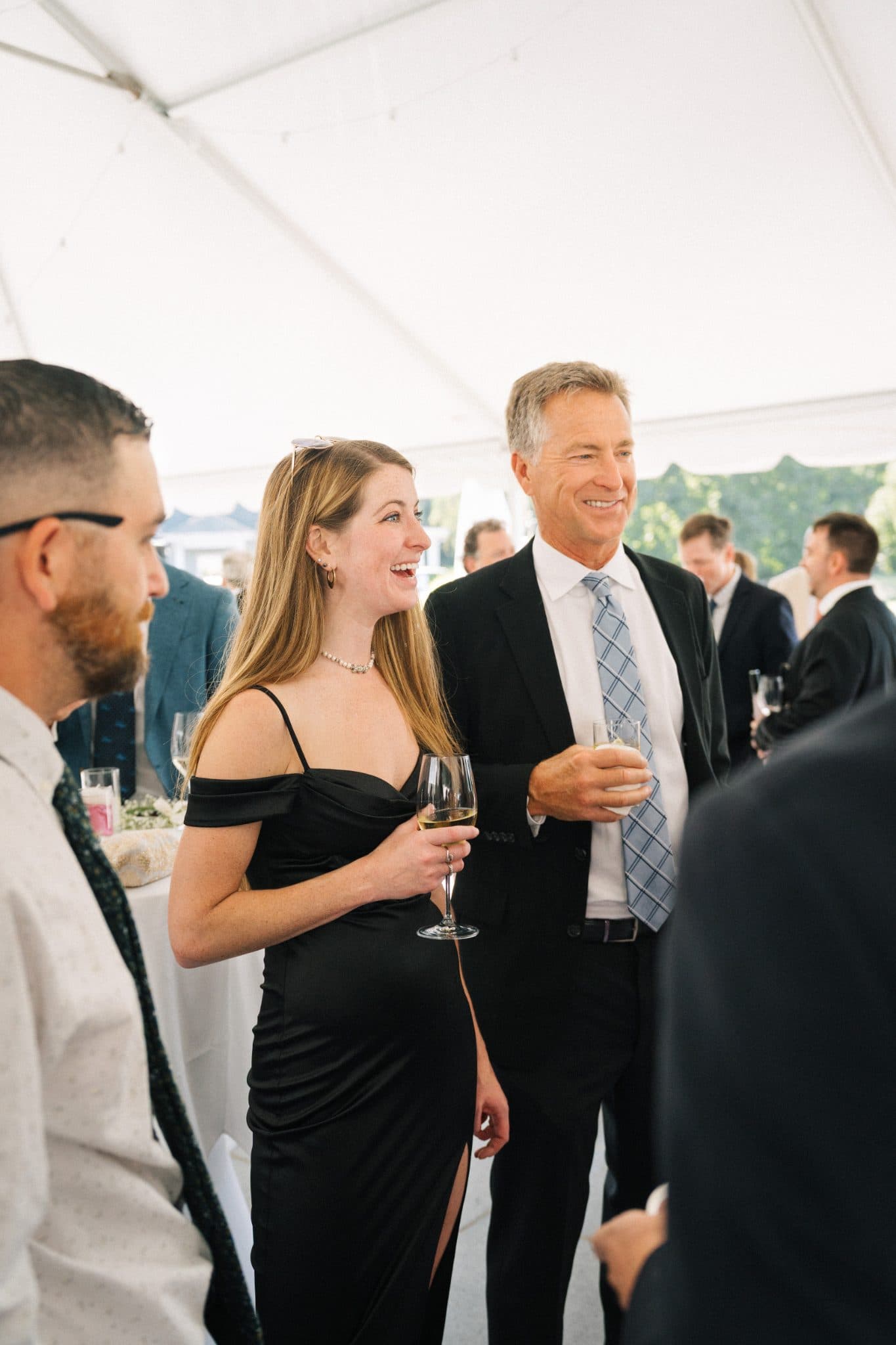 A woman in a black dress and a man in a suit are smiling and holding drinks under a tent.