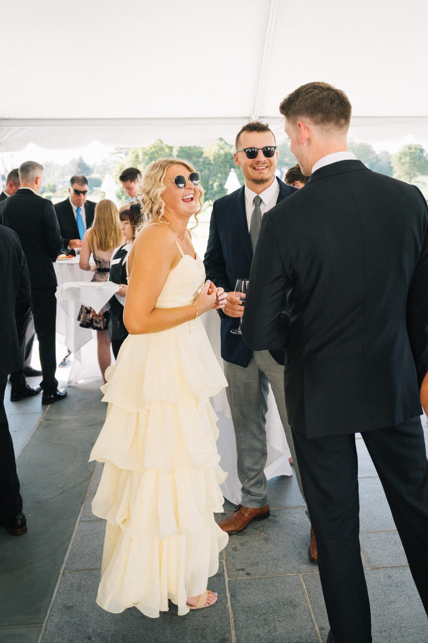 A woman in a tiered cream dress smiles while conversing with two men in suits at a wedding reception.
