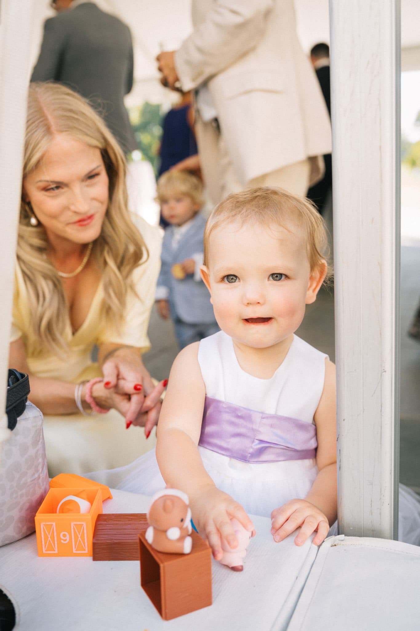 A baby in a white dress with a purple sash plays with toys on a table while a woman in a yellow top looks on.