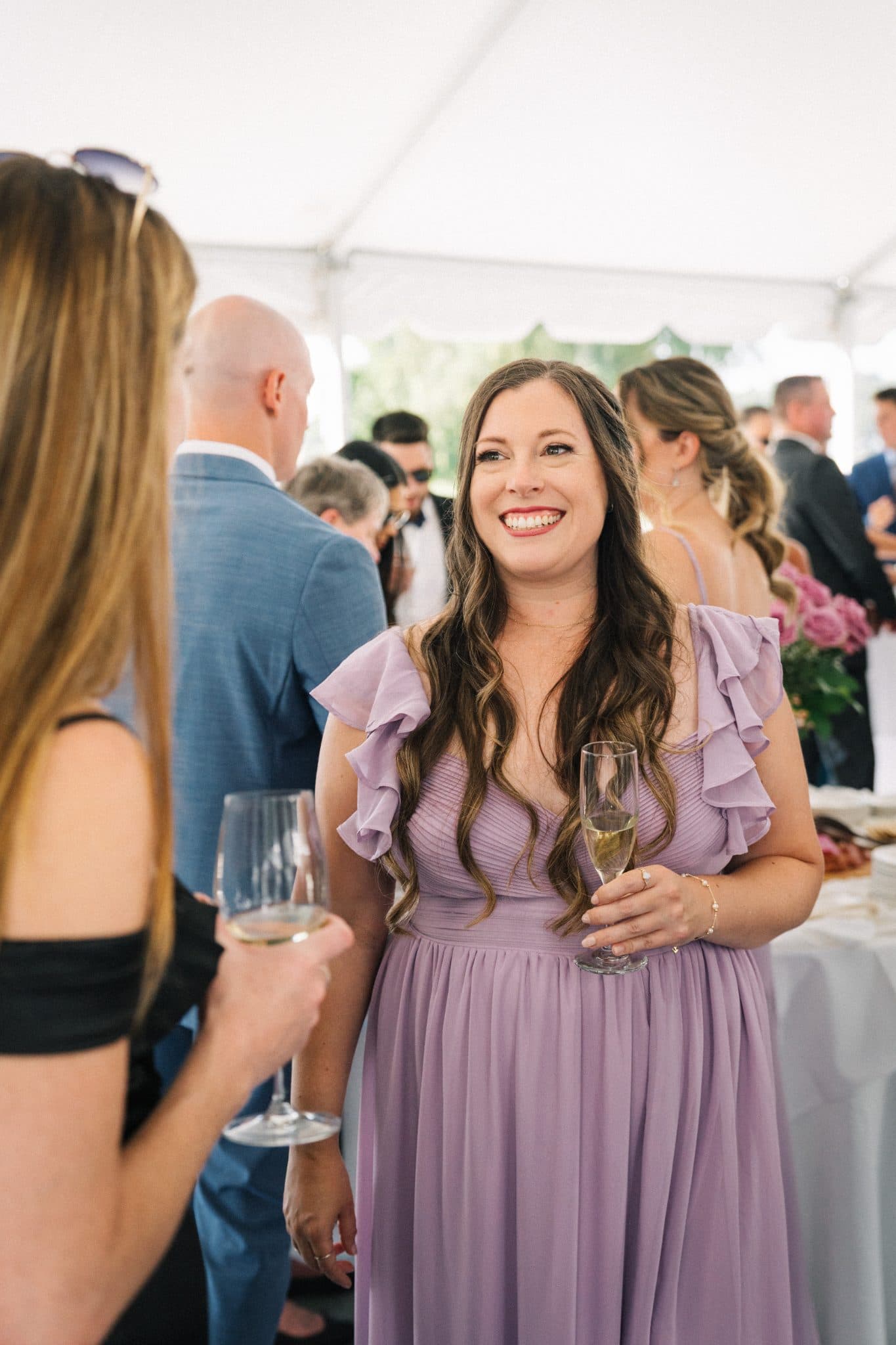 A woman in a lavender dress smiles while holding a glass of champagne at a wedding reception.