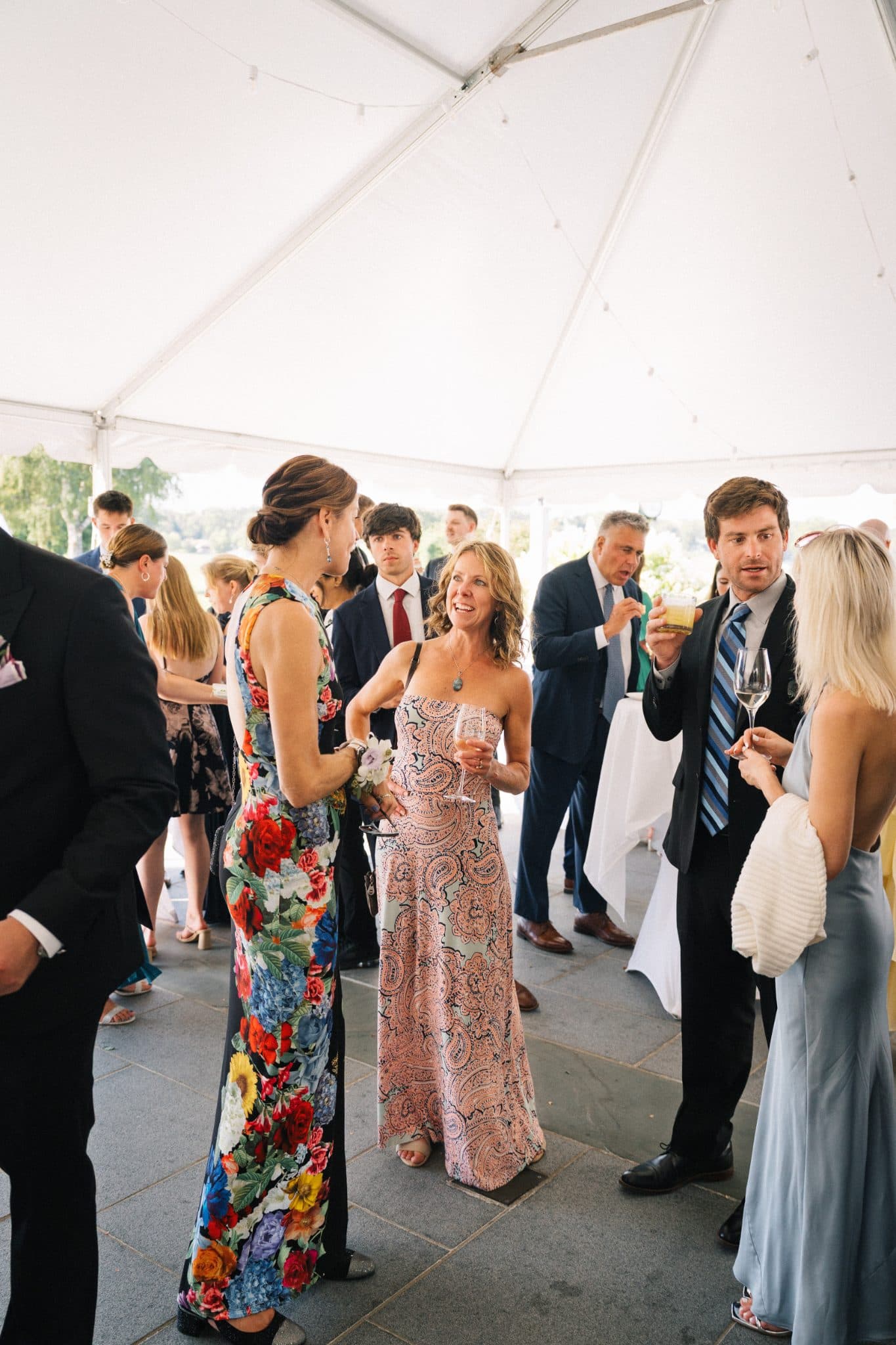 A group of people in formal attire stands under a white tent, holding drinks and engaging in conversation.