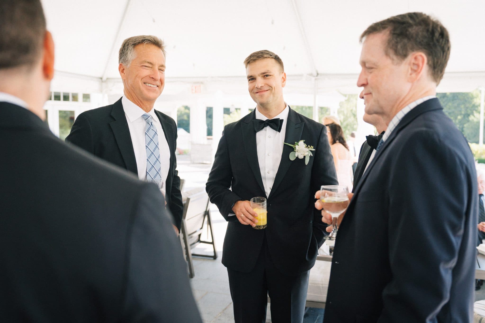 Three men in formal attire stand together, smiling, with drinks in hand, under a white tent.