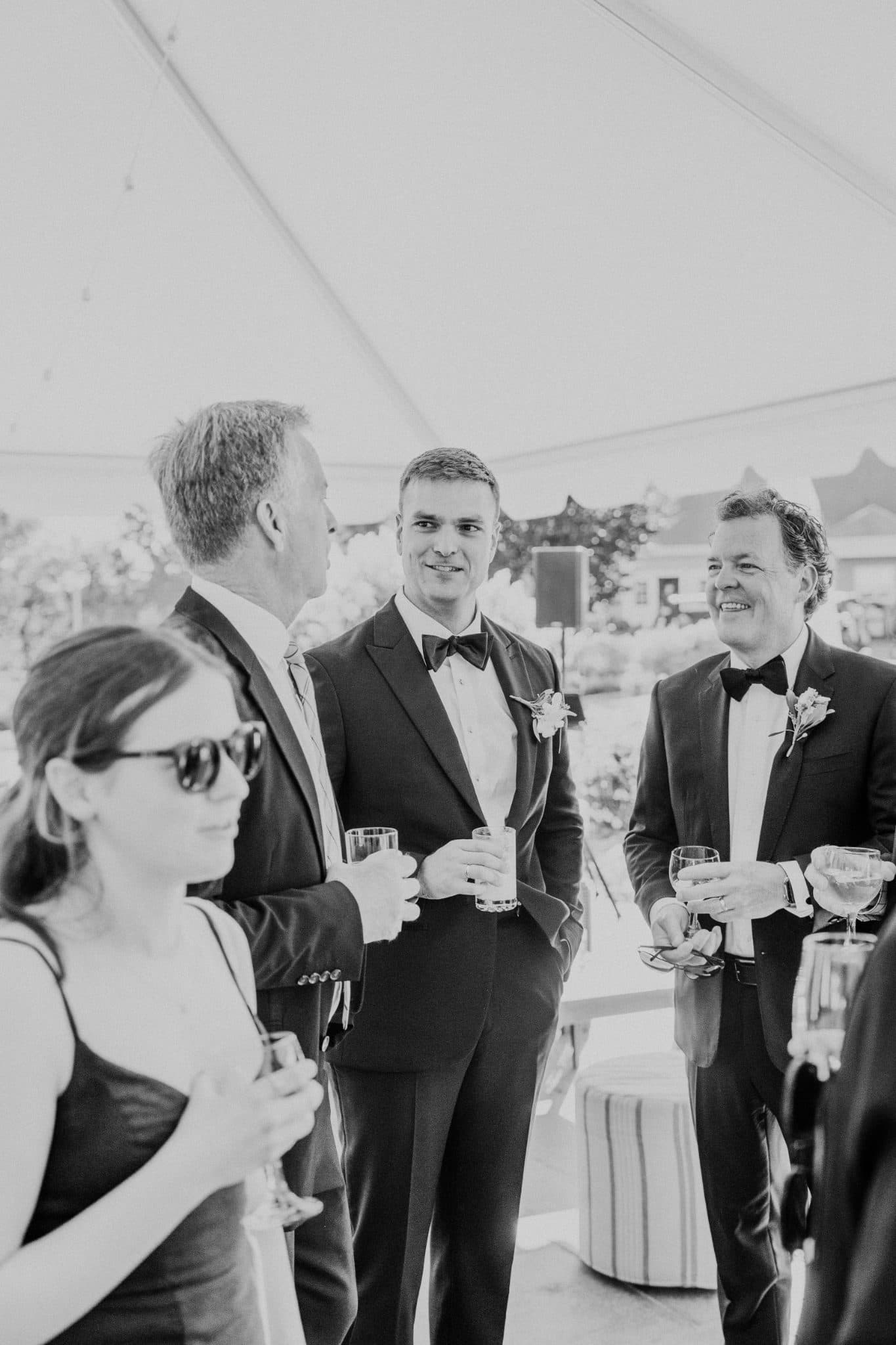 A group of people in formal attire stands together, holding drinks, under a tent at an outdoor event.