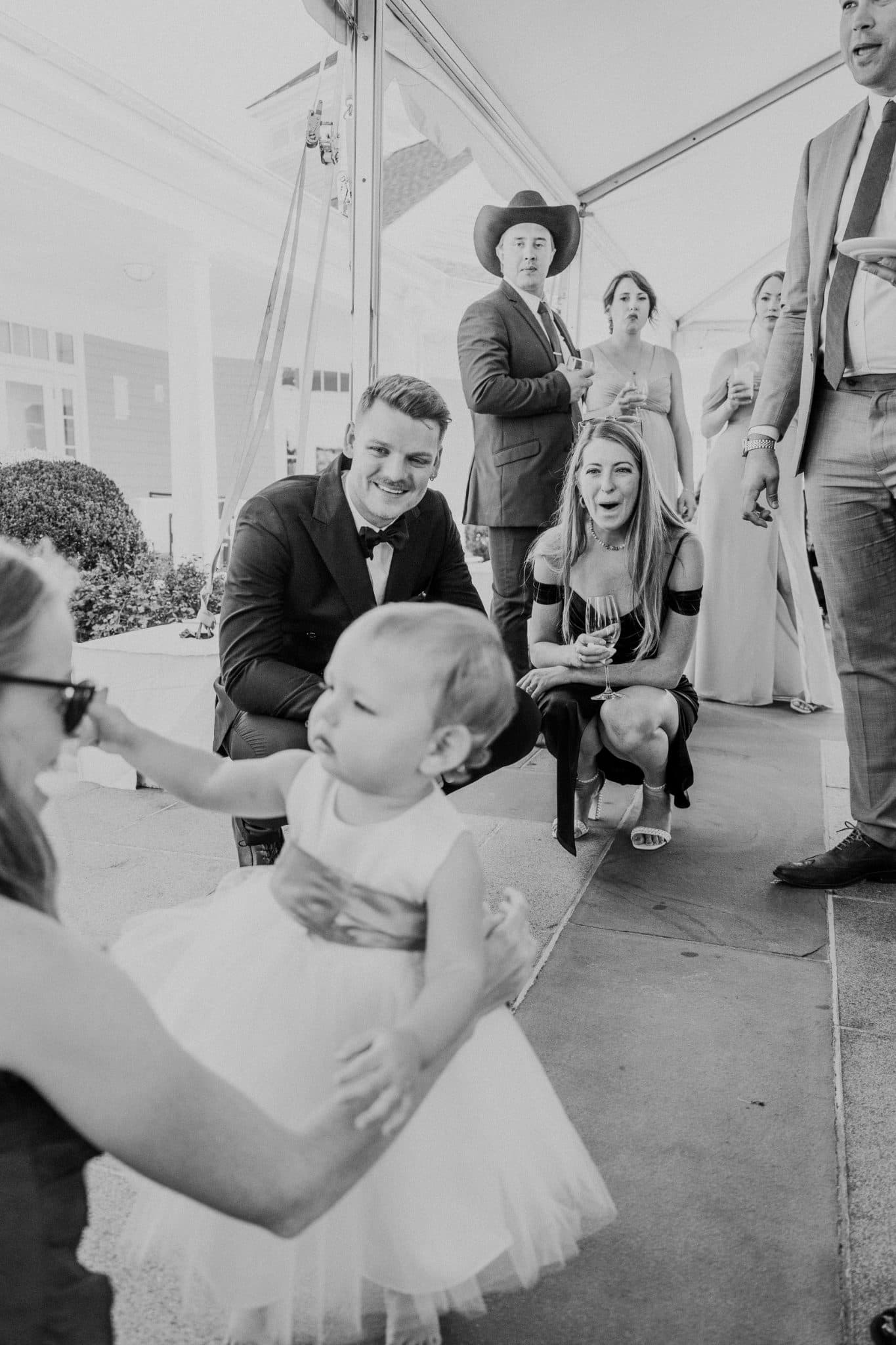 A black-and-white wedding scene shows a baby in a tutu being held, with adults in formal attire and a woman in a dress holding a glass of champagne, all under a tent.