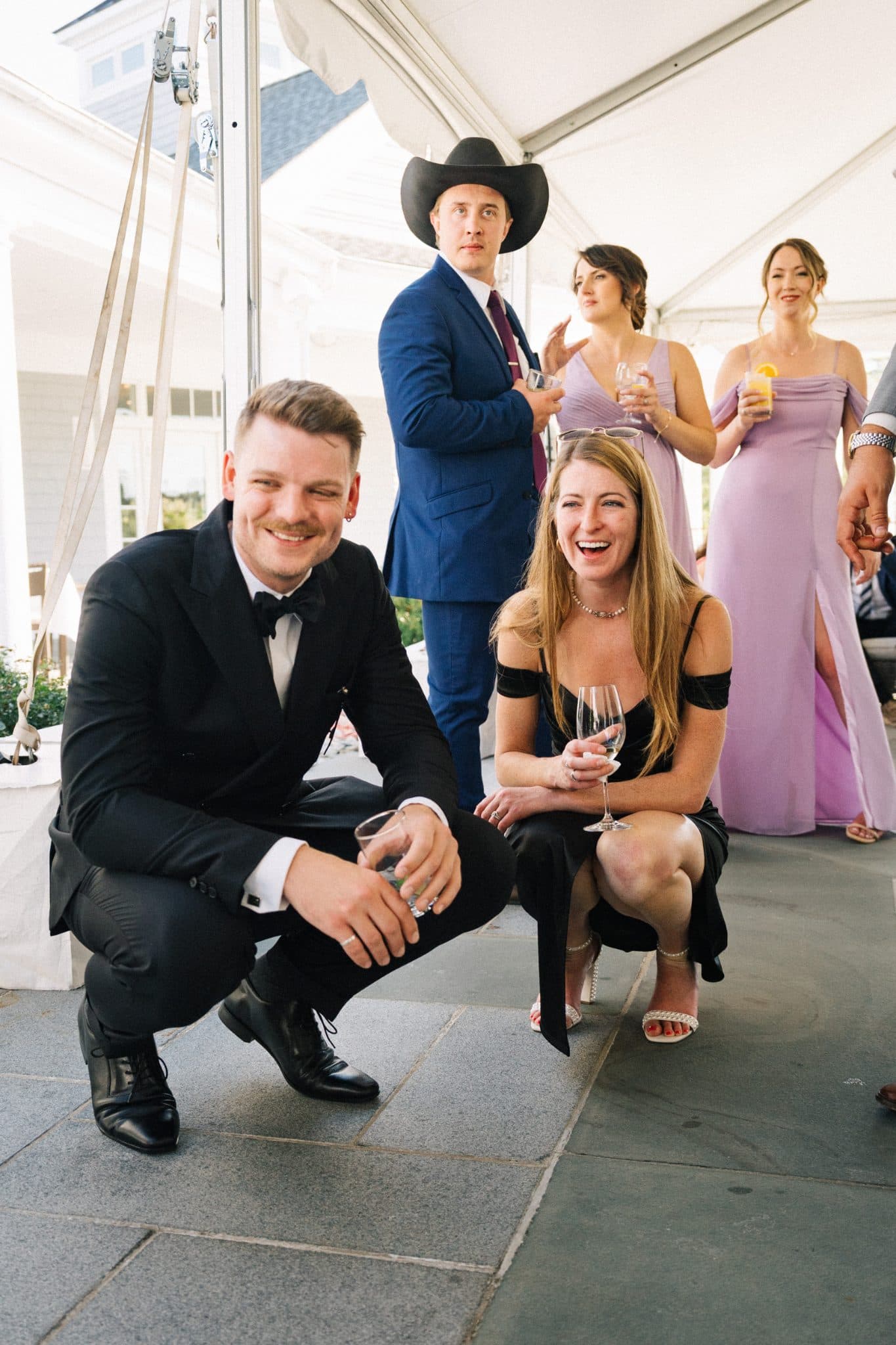 A man in a black suit and bow tie crouches next to a woman in a black dress holding a wine glass, with two women in lavender dresses and a man in…
