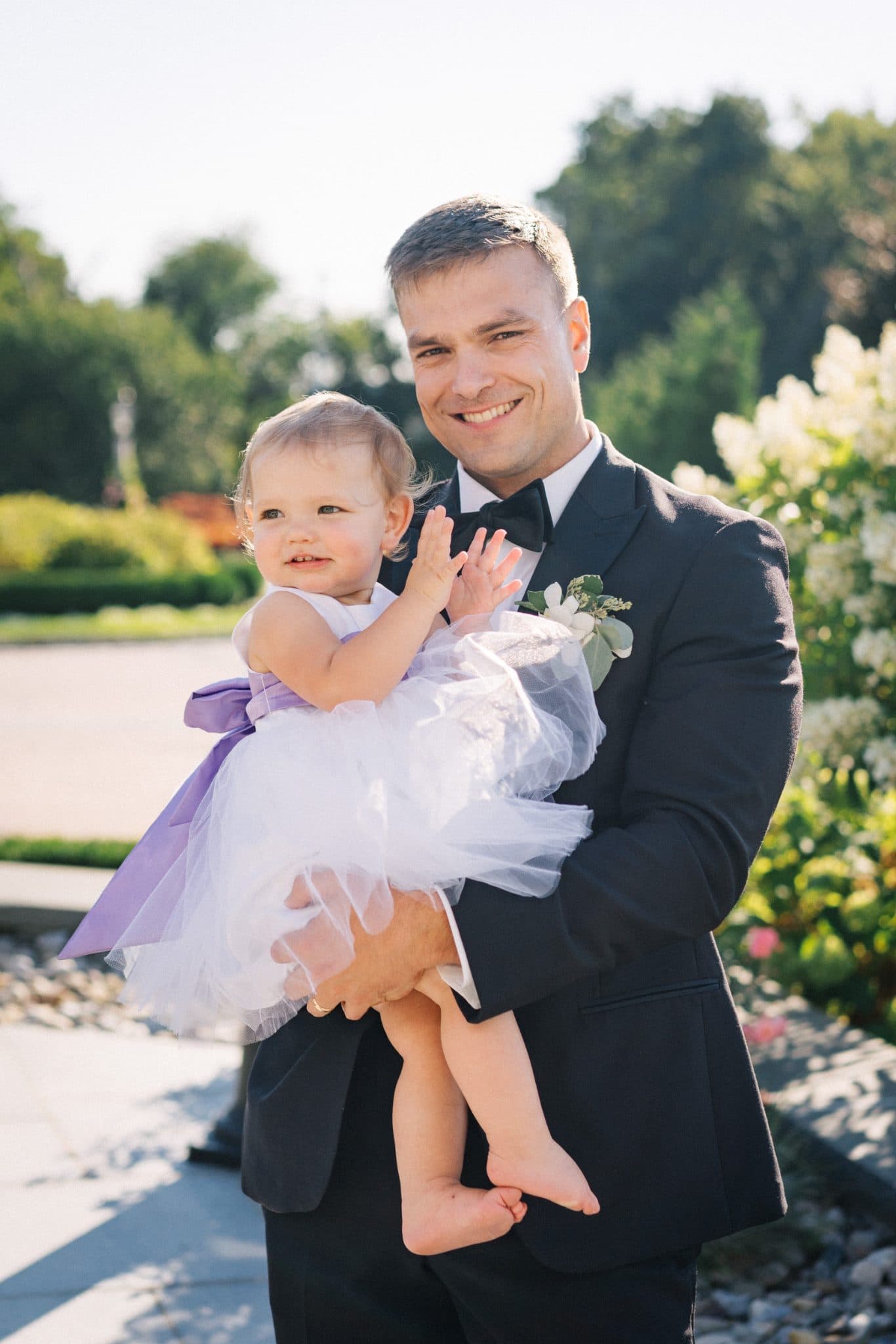 A man in a suit holds a baby dressed in white and purple, smiling under a clear sky with greenery in the background.