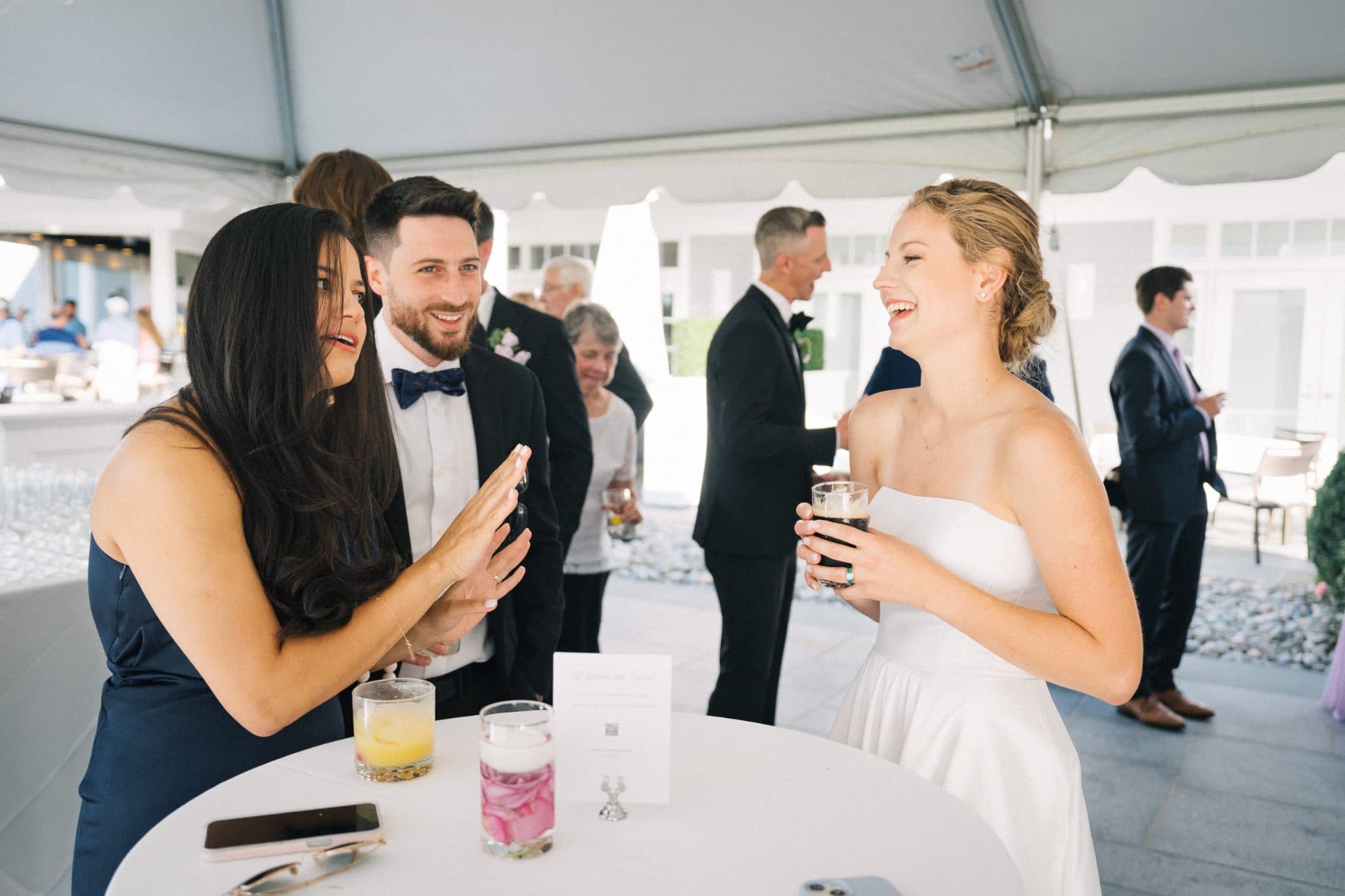 A bride in a white dress and a guest in a blue dress interact near a table with drinks and a card at a wedding reception.
