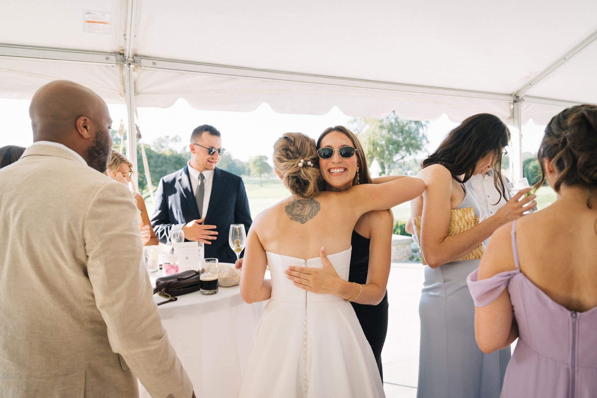 A bride in a white dress embraces a guest in a black dress under a white tent at a wedding reception.