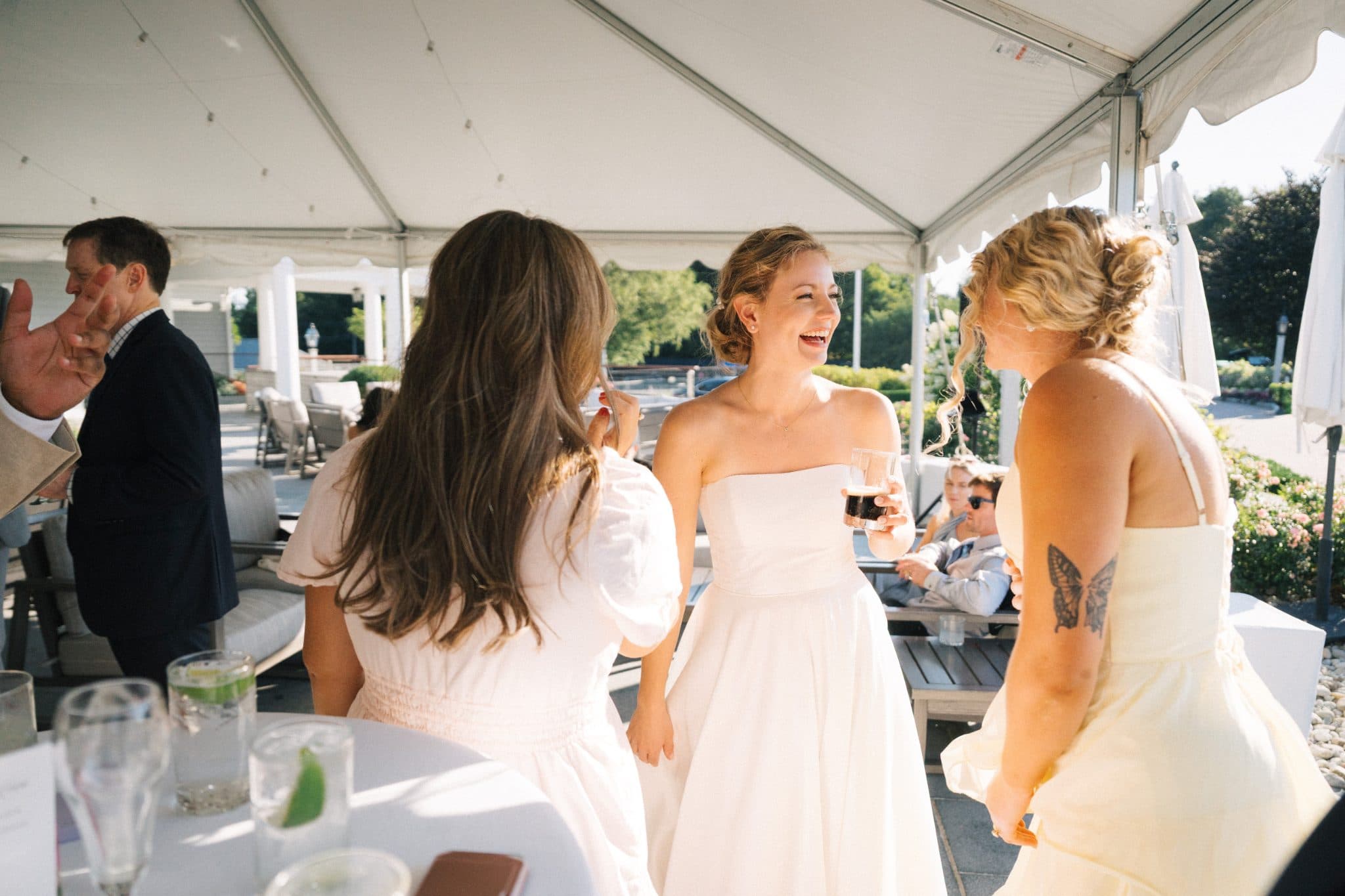 A bride in a white dress stands with two women in white dresses under a tent, holding drinks and smiling.