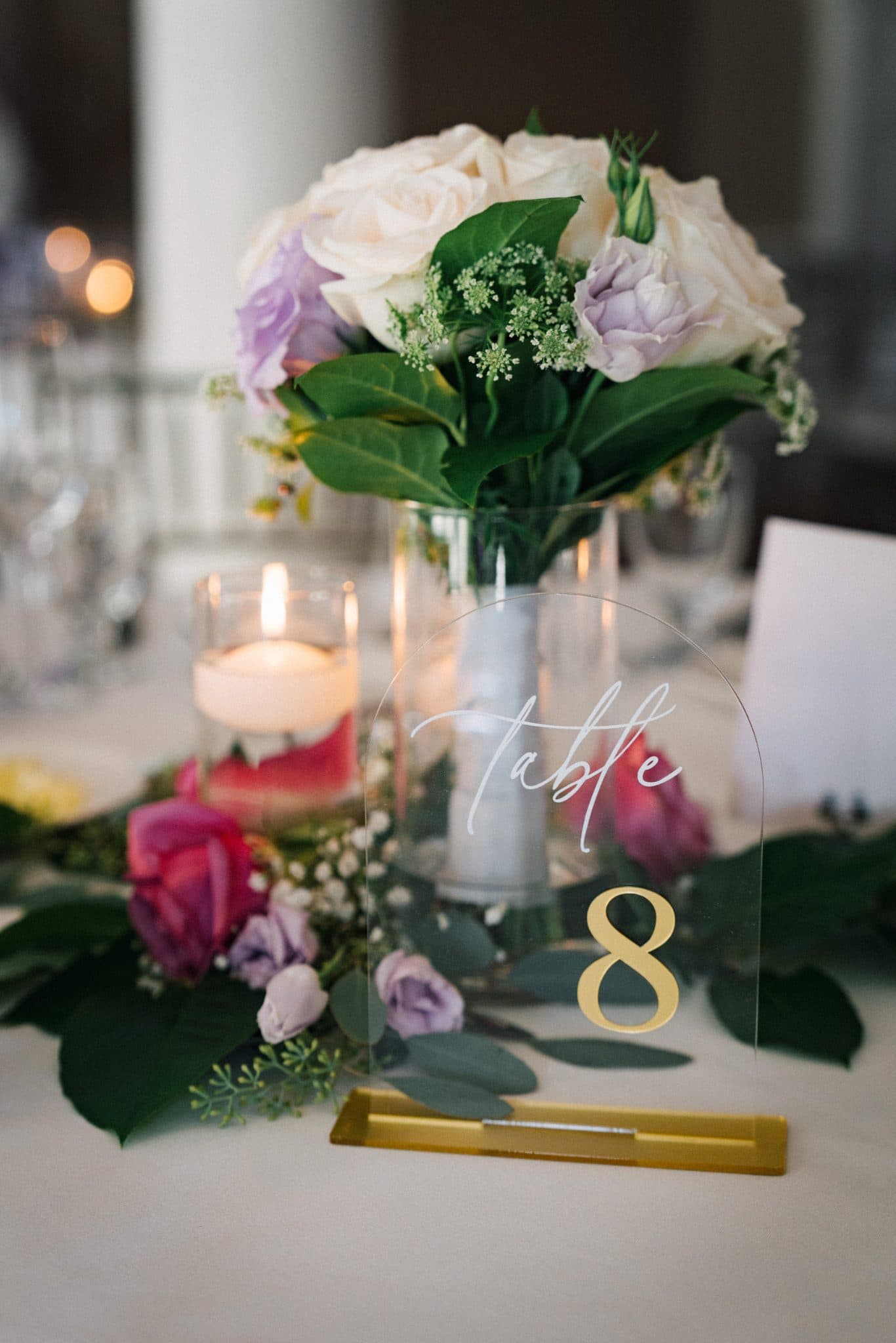 A table centerpiece features a vase with white and purple roses, greenery, and a lit candle, with a table number "8" in gold.