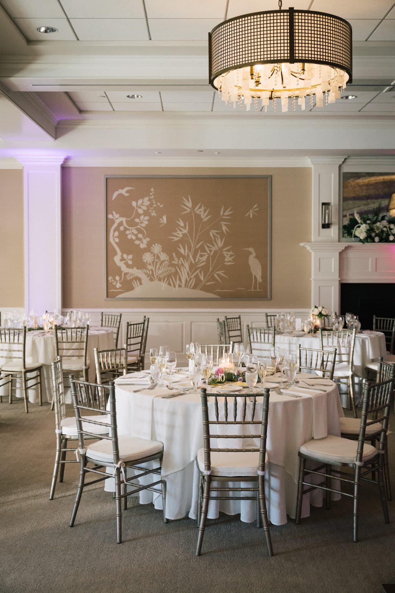 A warmly lit dining room with round tables, white tablecloths, silverware, and floral centerpieces, surrounded by chairs and a decorative wall mural.