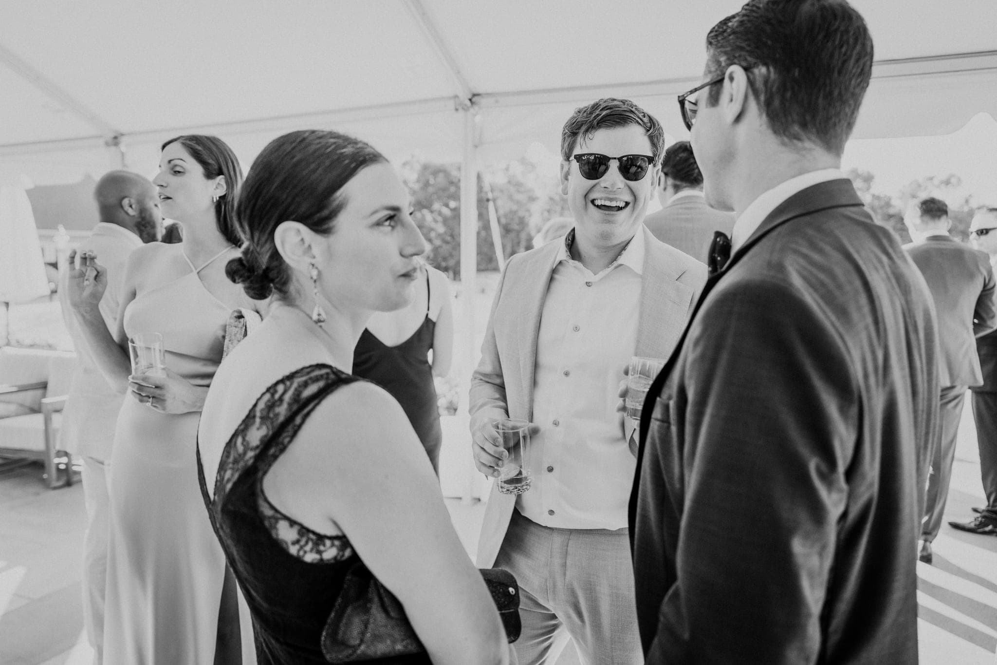 A black-and-white wedding photograph shows a group of people conversing under a tent, with one man in sunglasses smiling and holding a drink.