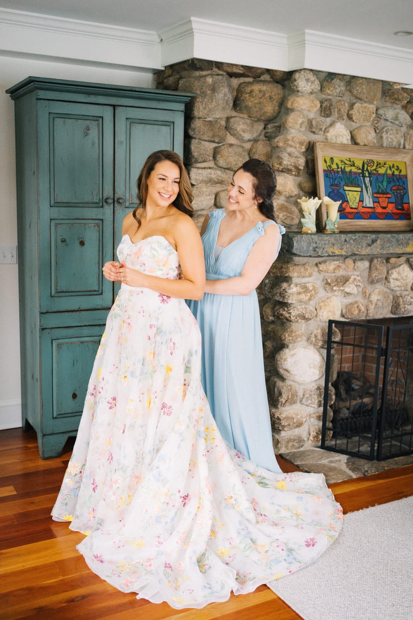 Two women in elegant dresses stand near a stone fireplace and a teal cabinet.