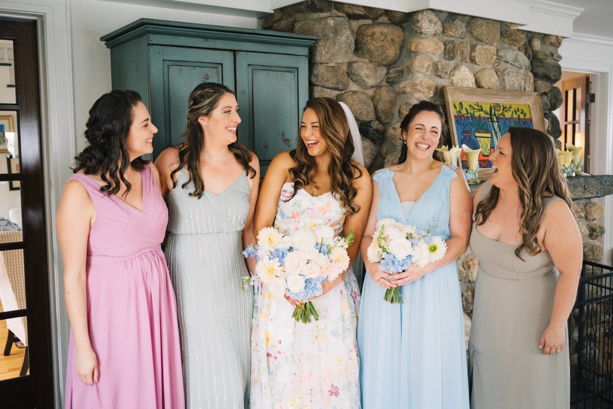 Five women in pastel dresses stand together, holding bouquets, smiling in front of a stone fireplace.