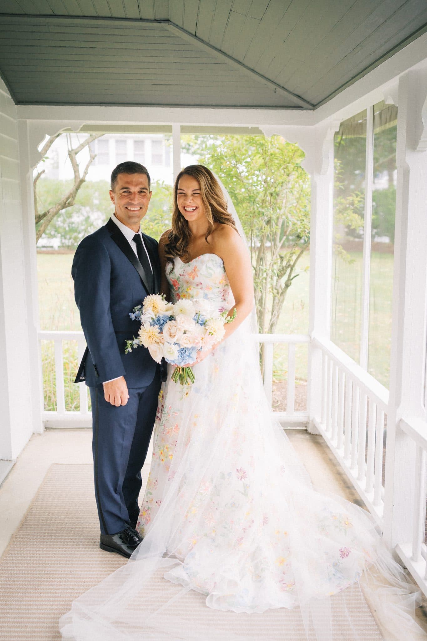 A bride in a floral wedding dress and a groom in a dark suit stand on a porch, holding a bouquet of flowers.