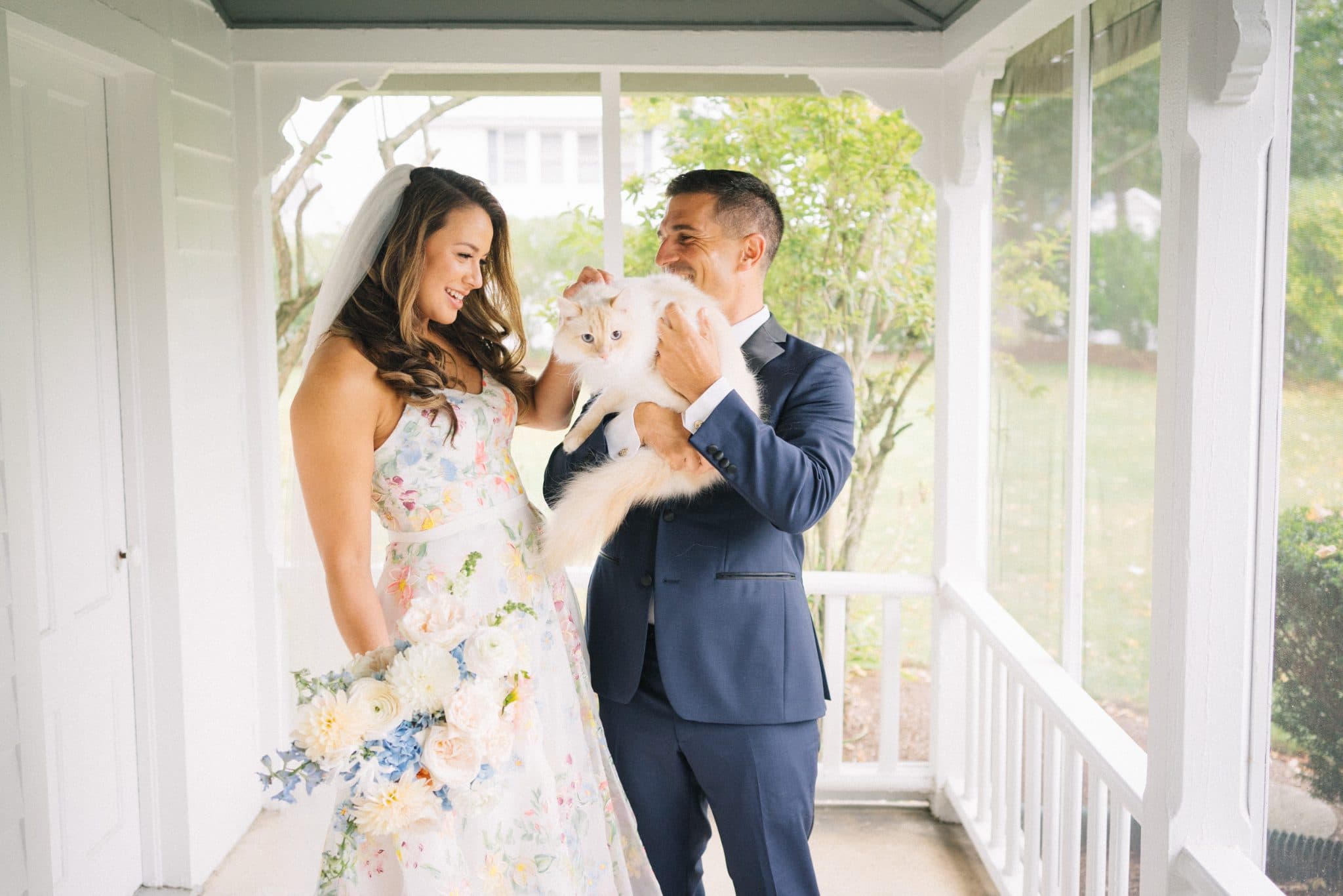 A bride in a floral dress and a groom in a blue suit hold a fluffy white cat on a porch with white railings.