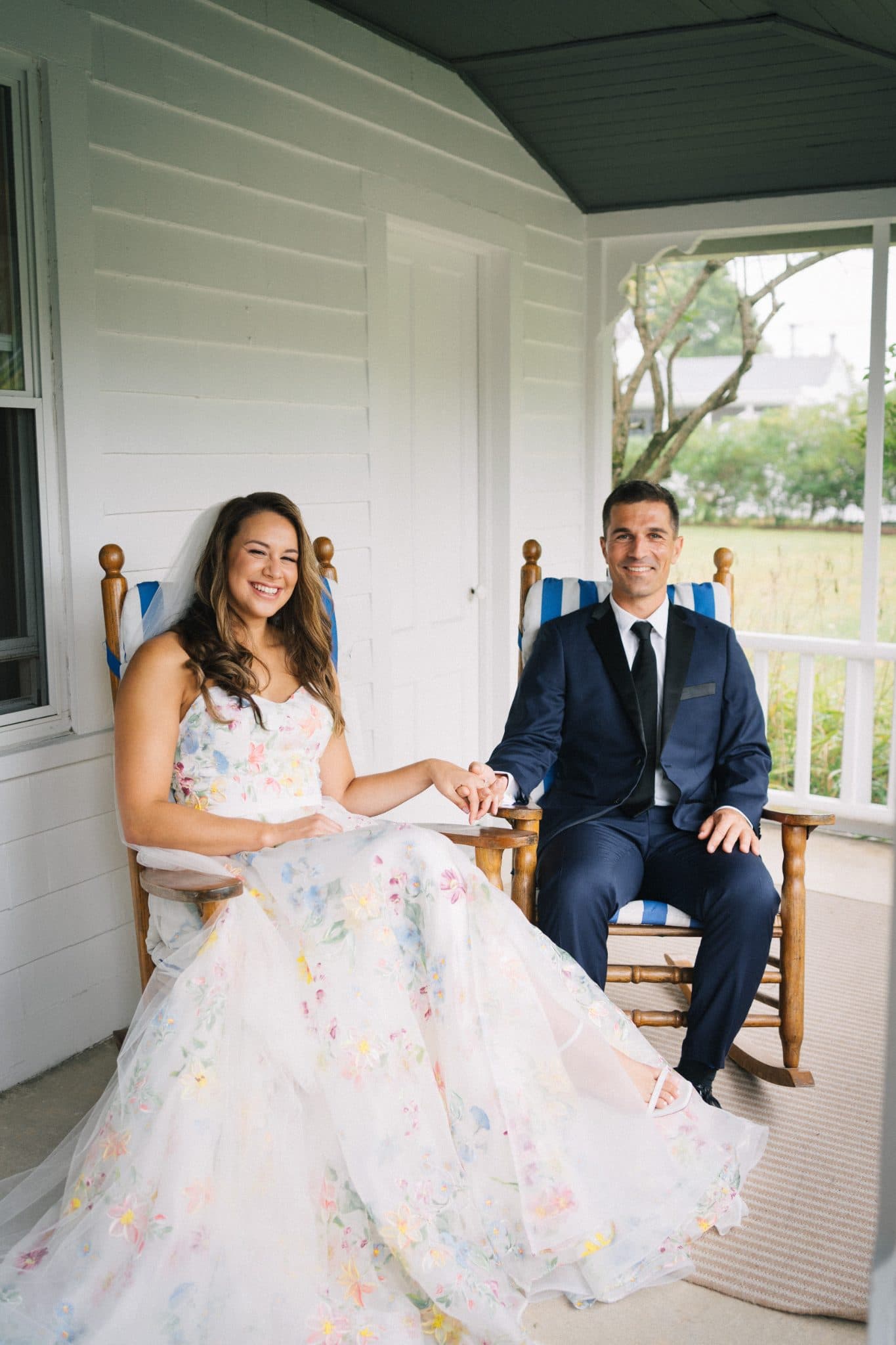 A couple sits on a porch, the woman in a floral dress and the man in a suit, both smiling.