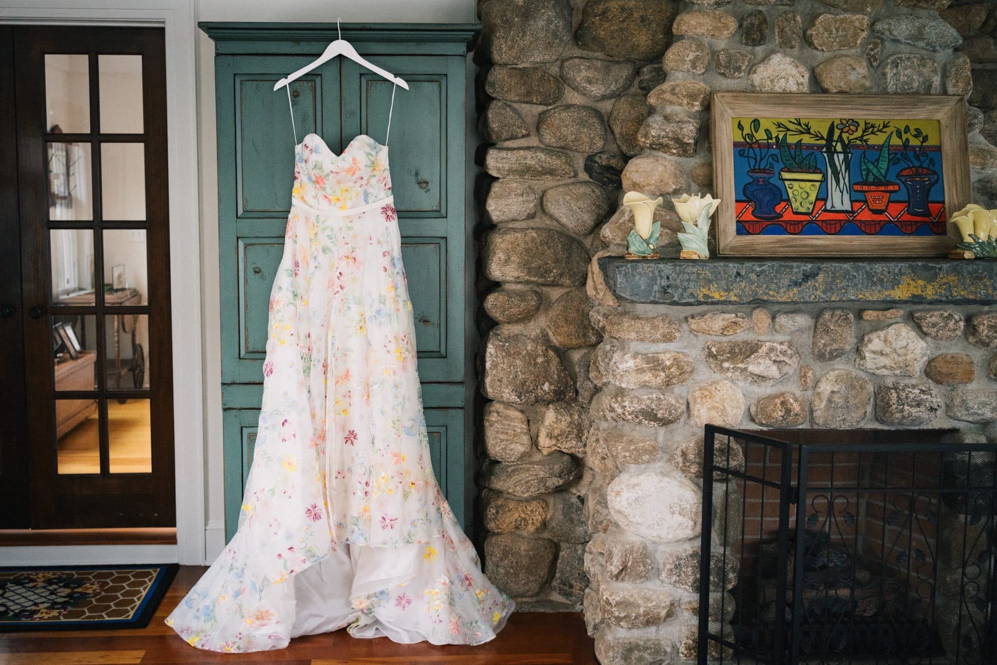 A floral wedding dress hangs on a hanger in front of a green door and a stone fireplace with decorative items.
