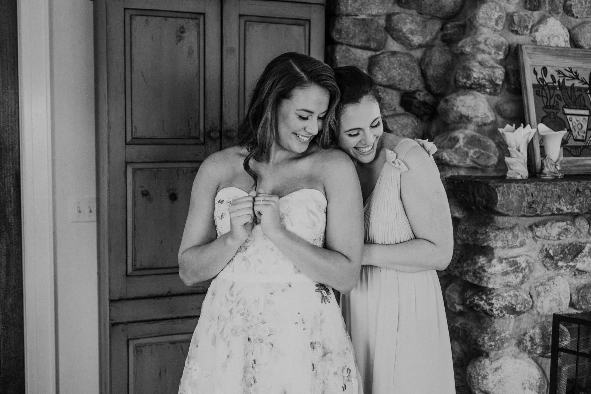 Two women, one in a strapless white dress, stand close, smiling, in front of a stone wall and wooden door.