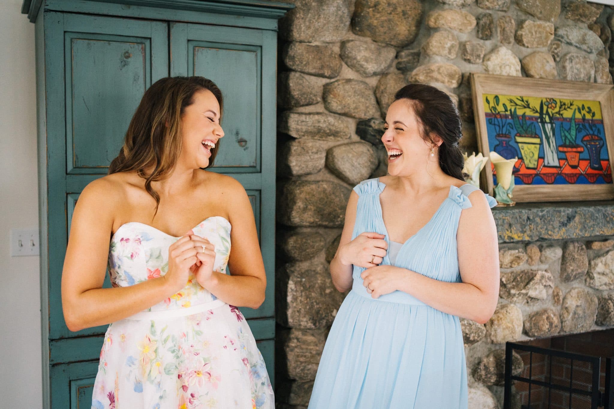 Two women in dresses laugh together in front of a stone fireplace and teal cabinet.