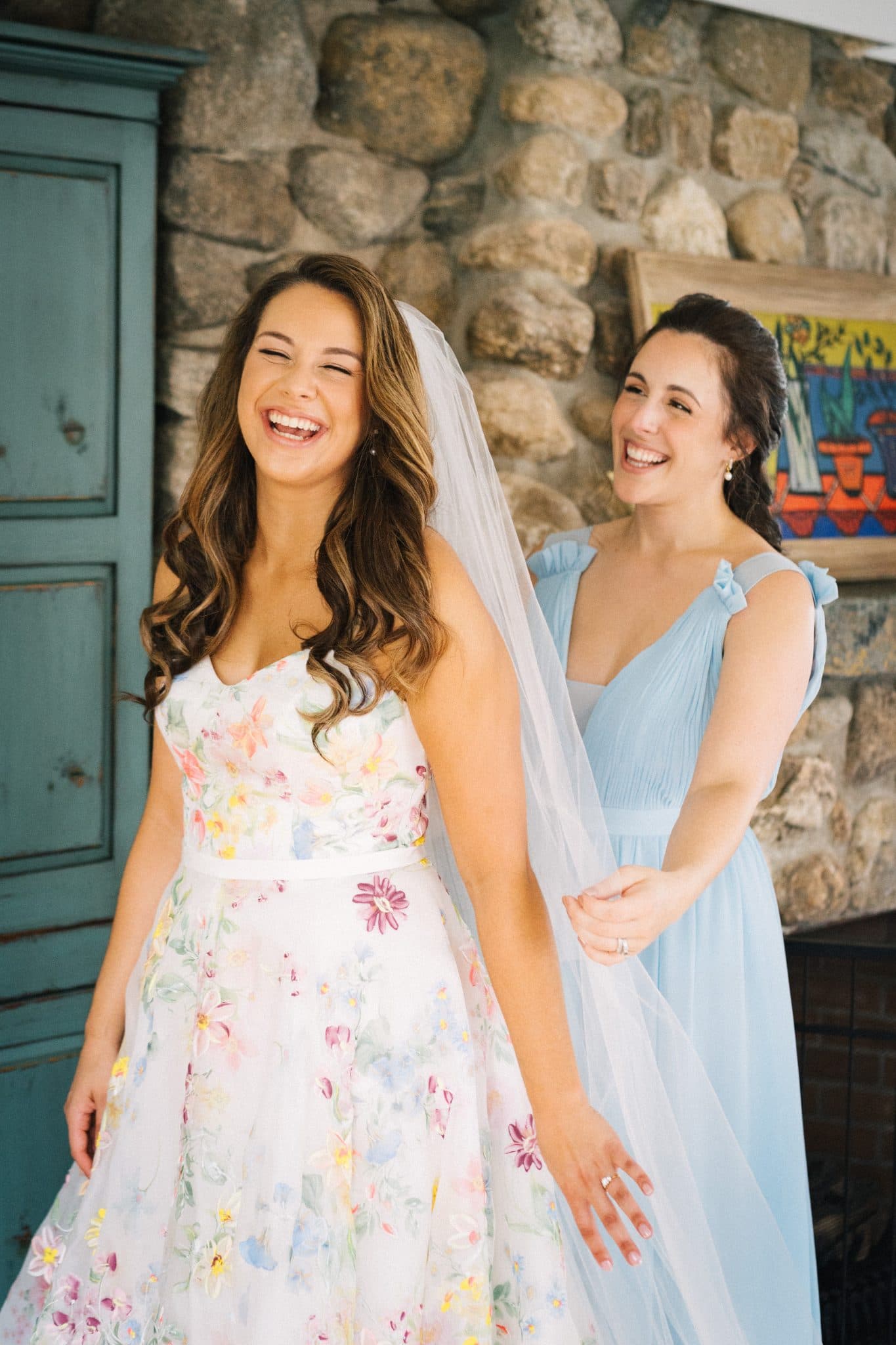 Two women in dresses, one in a floral dress with a veil, smiling in front of a stone wall.