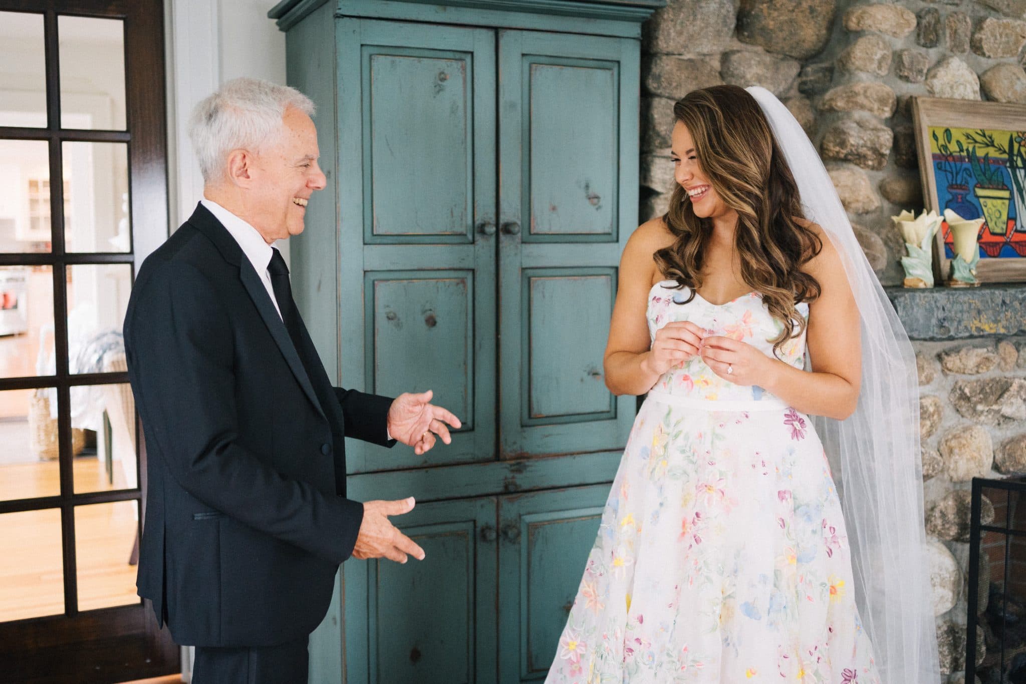 A bride in a floral dress and veil stands beside a man in a suit, both smiling and gesturing towards each other.